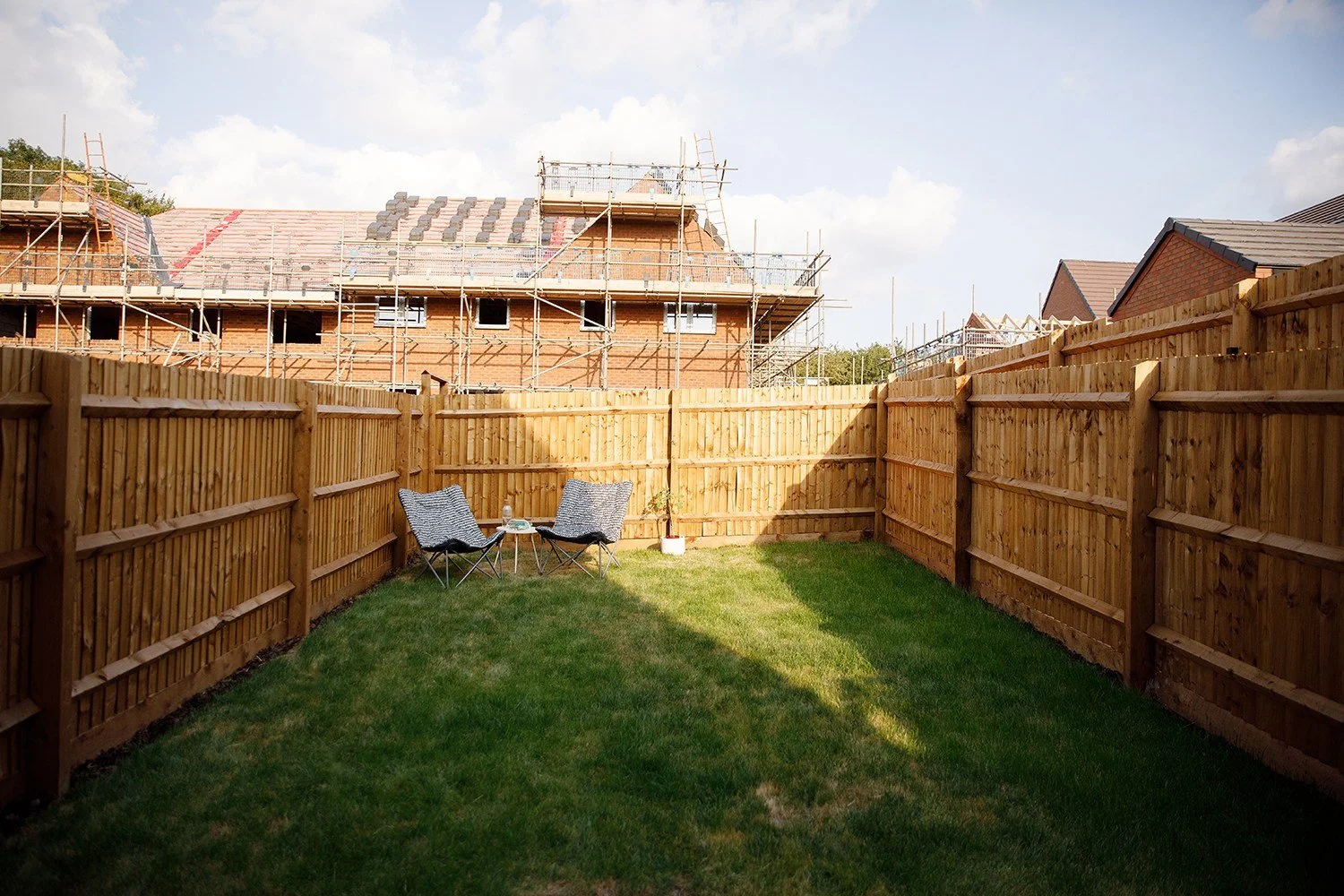 Backyard with green grass, wooden fence, two outdoor chairs and a small table, construction site with scaffolding and brick house in the background.