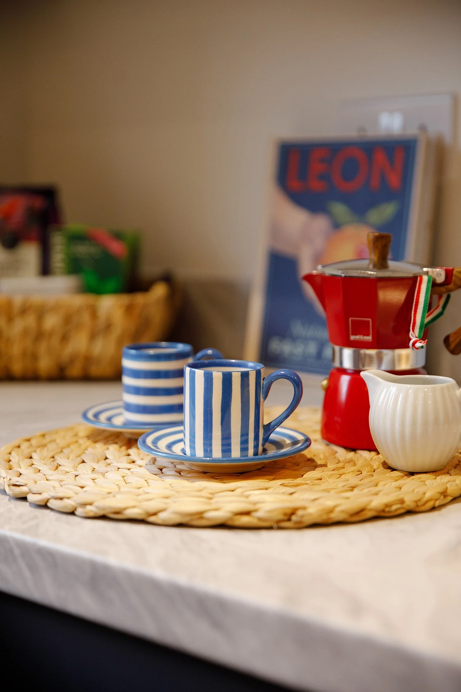 Set of blue and white striped coffee cups and saucers on a woven placemat, with a red moka pot and white creamer pitcher, in a kitchen with a basket of assorted products and a poster in the background.