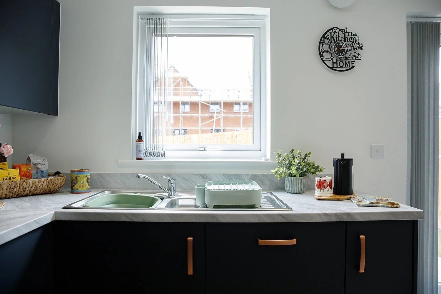 Kitchen with black cabinets, a marble countertop, and a window above the sink. The sink has a chrome faucet, a dish drying rack, and a small potted plant on the counter. A circular wall clock with a decorative design reads 'The kitchen is the heart o