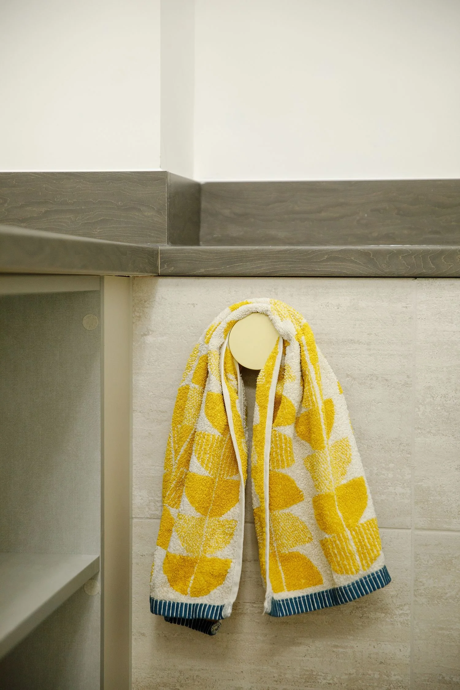 Yellow and white striped towel hanging on a beige hook on a light-colored wall in a bathroom or locker room, with part of a wooden or laminate shelf visible on the left.
