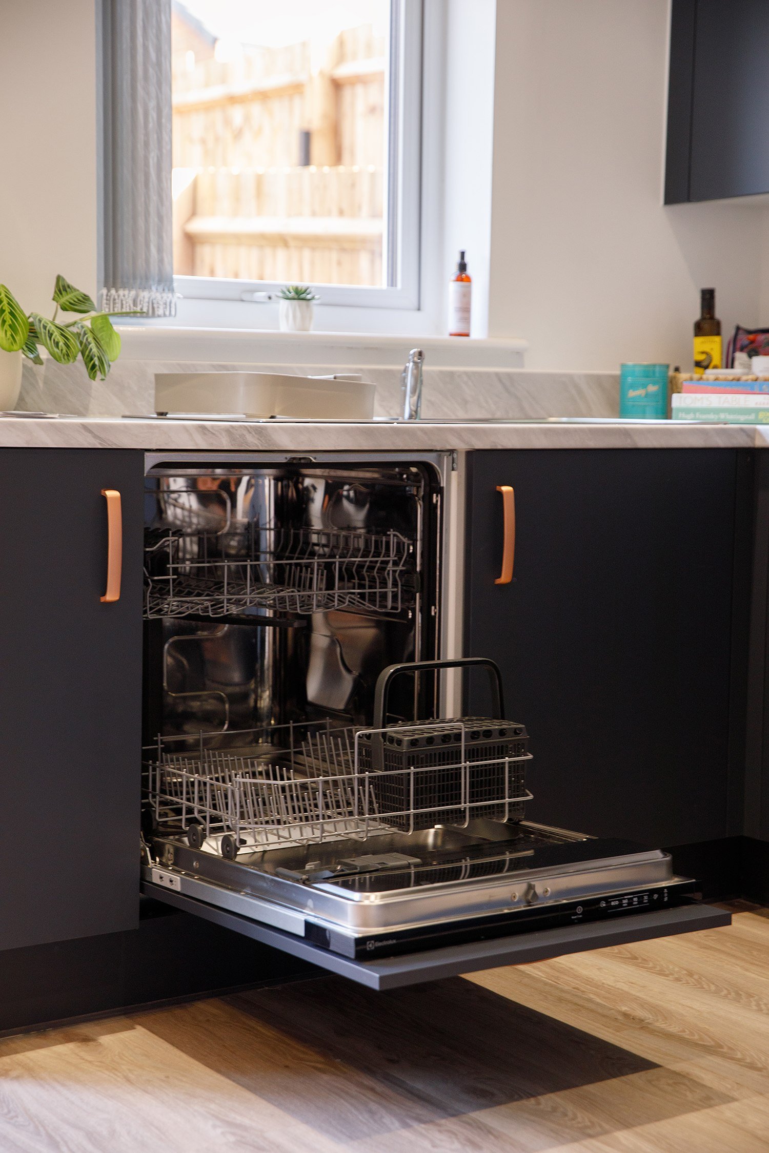 Open dishwasher with racks inside, kitchen counter with plants, bottles, and books nearby, window in the background, and wooden floor.