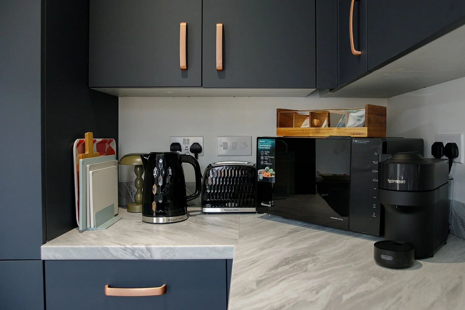 Kitchen countertop with black kettle, toaster, microwave, Nespresso machine, and wooden spice rack, with dark cabinets above and a light-colored countertop.