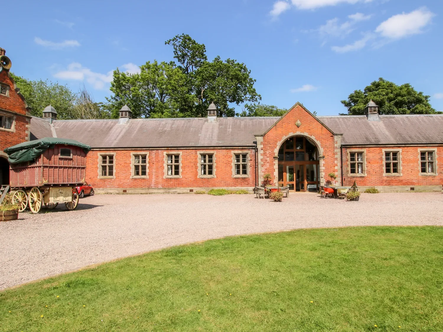 A brick building with arched entrance, surrounded by outdoor seating and a vintage wagon, under a blue sky with trees in the background.