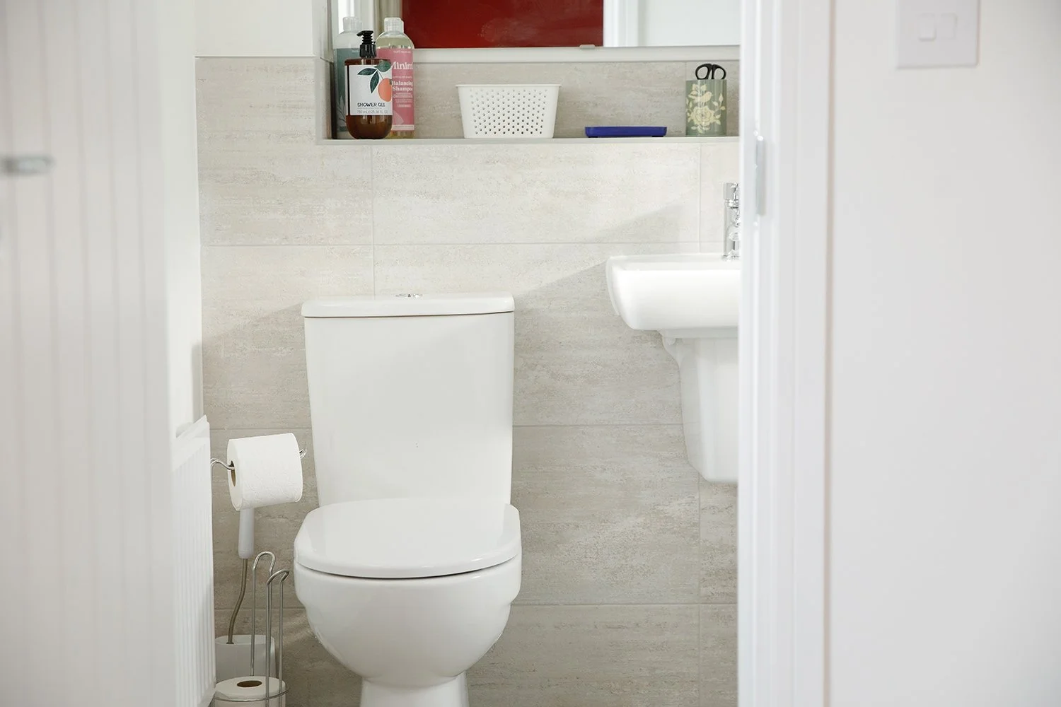 Bathroom with a white toilet, a small white sink, and a shelf above with toiletries, a basket, and a decorative box.