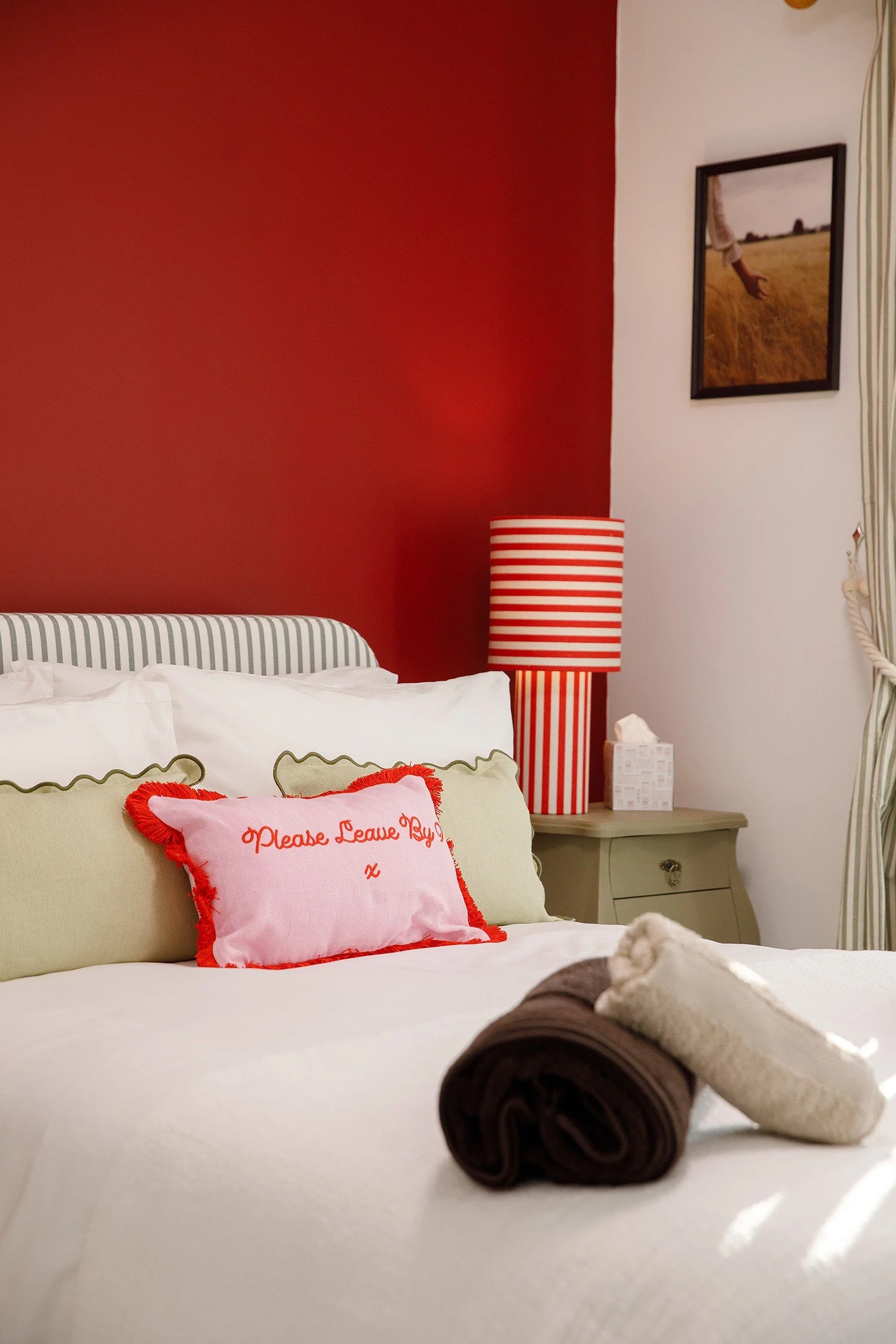 A neatly made bed with pillows, including a pink pillow with red fringes and embroidered text that says 'Please Leave Boy ...', and two rolled towels, one white and one dark brown, placed on the white bedspread. In the background, a bedside table wit