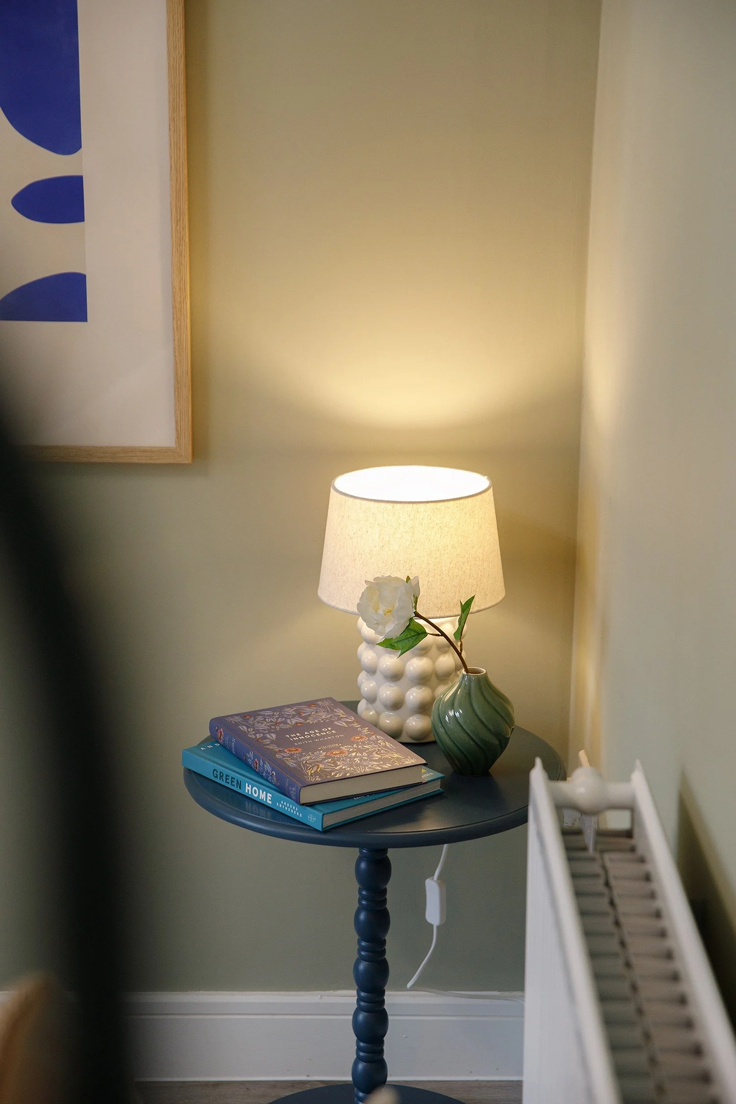 A round dark blue side table with a stack of three books on top, a white table lamp with a white orchid flower beside it, a green decorative vase, set against a pale yellow wall, with a painting partially visible on the left side.