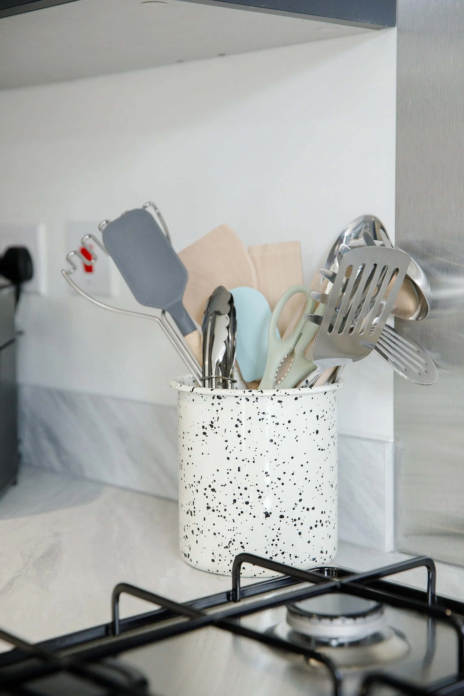 A kitchen countertop with a speckled white and black utensil holder containing various cooking tools, including a gray spatula, wooden spoons, a green and white spatula, and a metal slotted spatula. A gas stove with black grates is in the foreground,