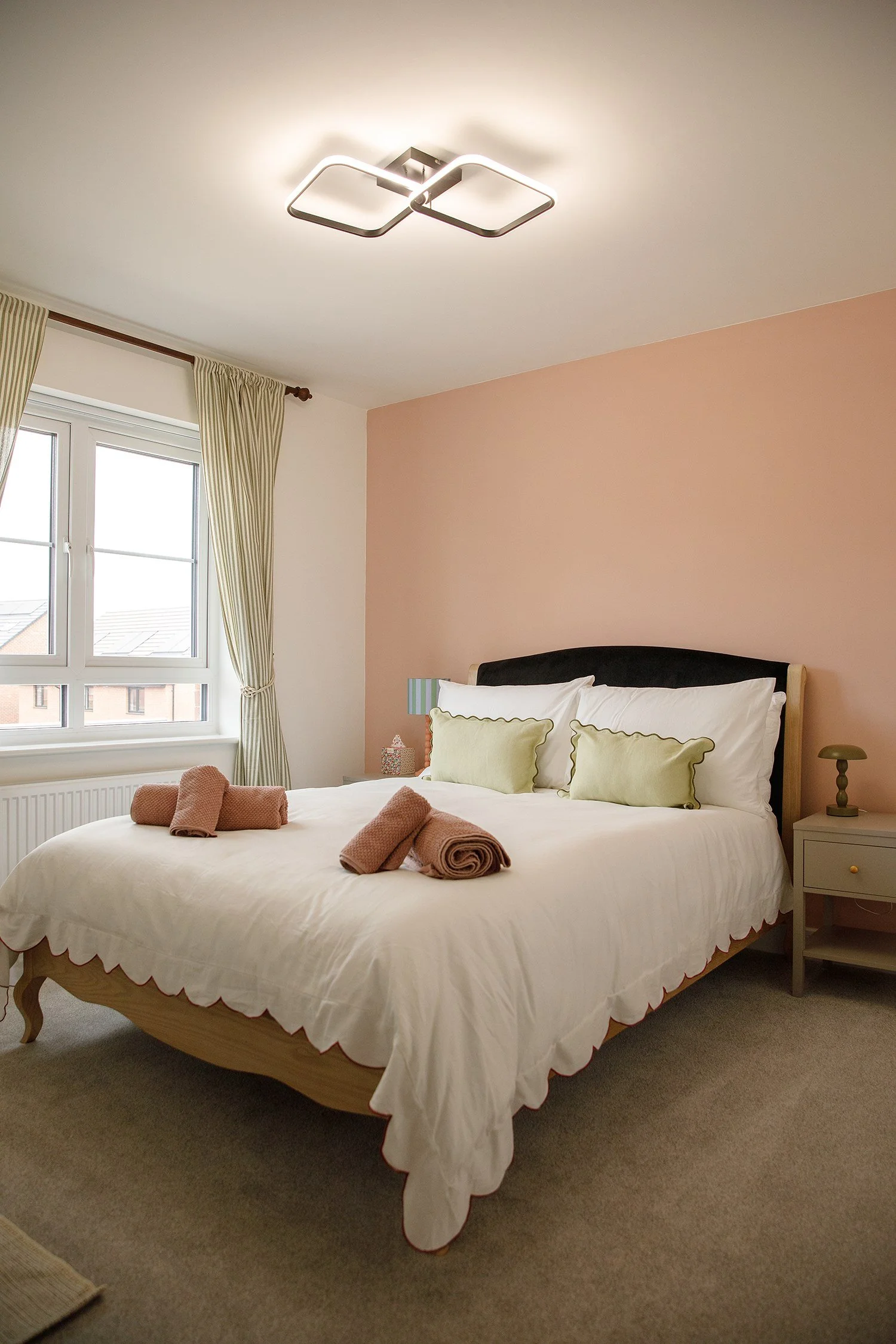 A bedroom with a large bed covered in white bedding, adorned with three light green pillows and four rolled brown towels. Next to the bed is a white nightstand with a small lamp, and the room has a window with cream-colored striped curtains. The wall