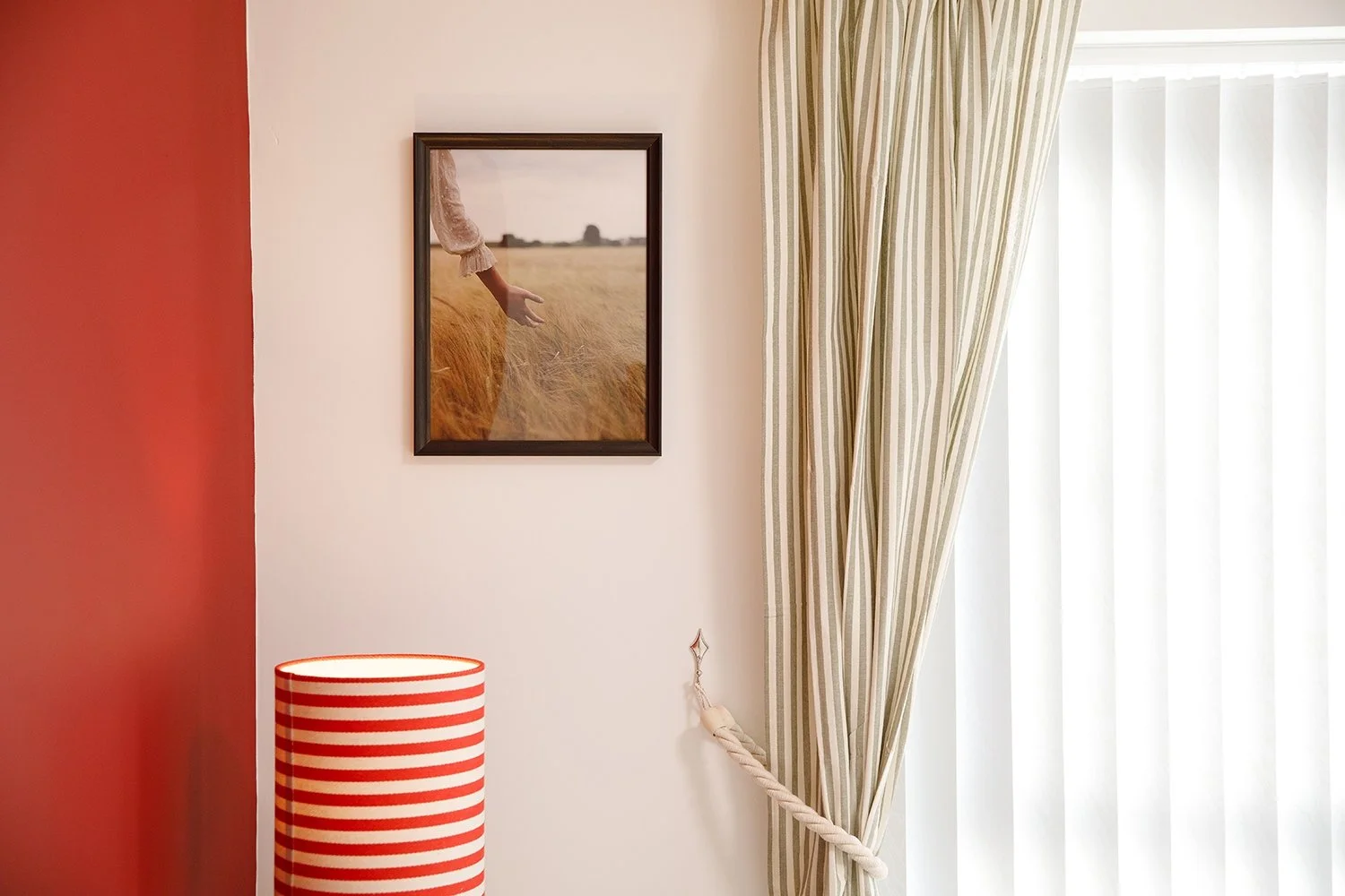 Interior part of a room with a red wall on the left, a white wall with a framed photo of a person in a field, striped curtains tied back on the right, and a red and white striped lampshade.