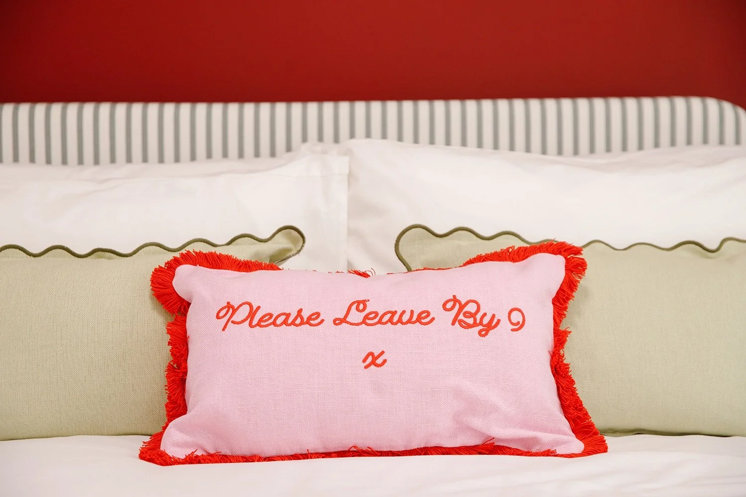 Decorative pink pillow with red fringe on a bed, embroidered with 'Please Leave By 9' in red script, in front of beige and white pillows with a striped headboard and red wall background.