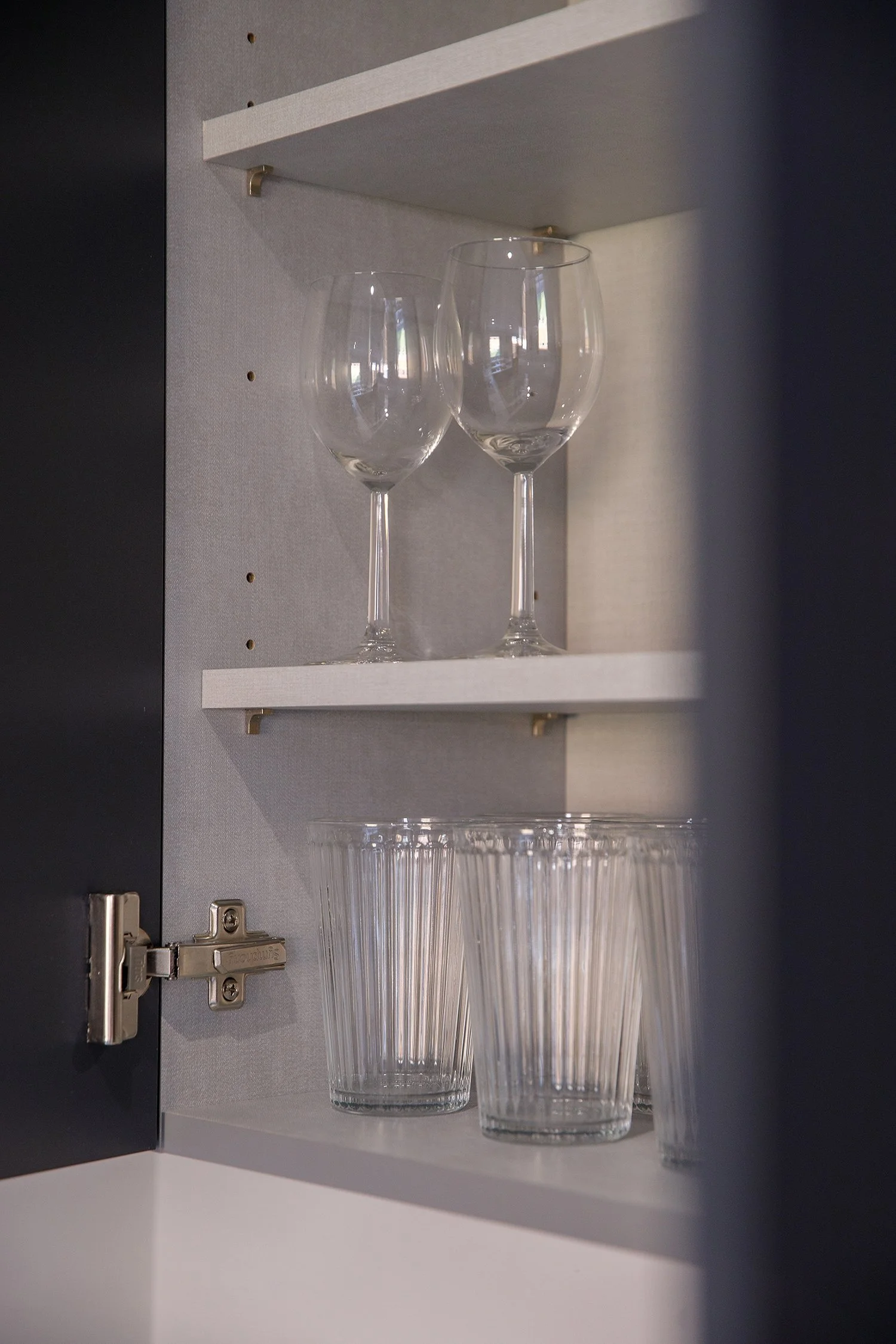 Two empty wine glasses and two empty tumbler glasses inside a cabinet with white shelves and a gray back panel.