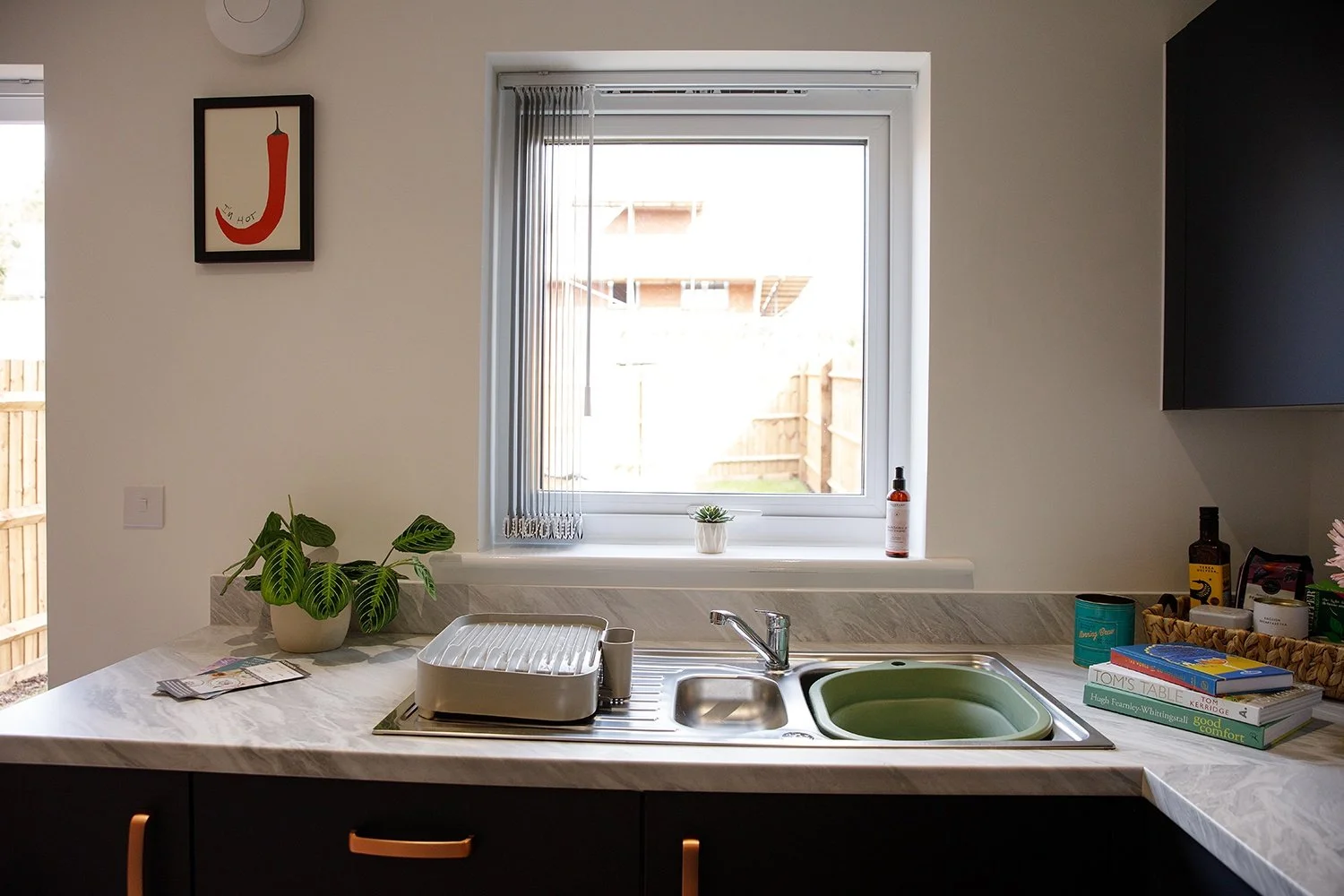 Kitchen sink with green basin, potted plant, window with blind, books, and small decorative items on the countertop.