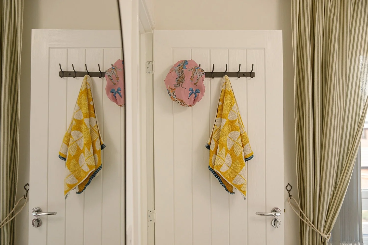 A door with a black hook rack holding two yellow towels with white and blue borders and a pink cloth with a tiger and blue bow pattern, reflected in a mirror, with striped curtains on either side.