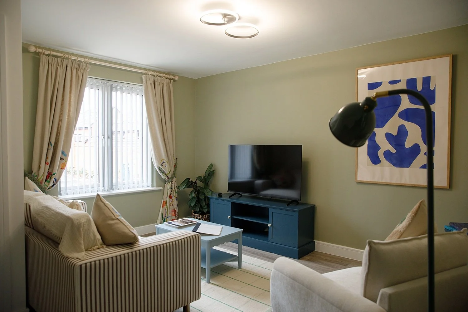 Living room with a striped sofa, a blue TV stand with a flat-screen TV, a coffee table with a book and a notebook, a plant, a window with cream curtains, and a large blue and white abstract art piece on the wall.