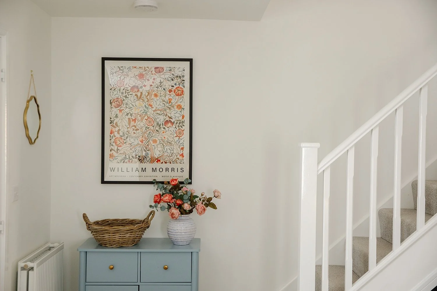 Interior of a home with framed artwork, a blue cabinet with a vase of pink flowers, a wicker basket, a wall mirror, and a staircase with beige carpeted steps.