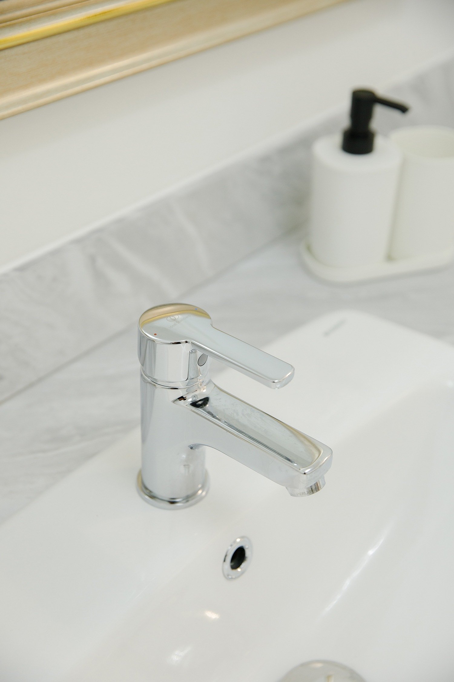 Close-up of a modern chrome bathroom faucet mounted on a white sink with a black soap dispenser and mirror in the background.