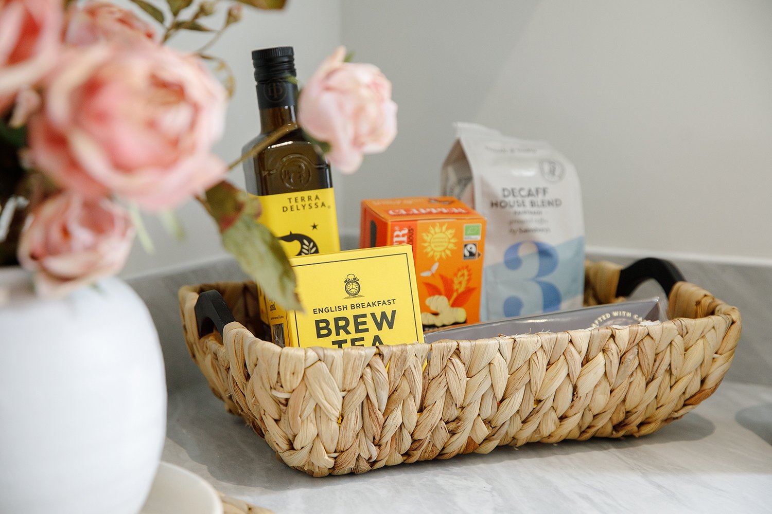 A woven basket containing tea, coffee, and syrup bottles on a countertop. Pink roses in a white vase are in the foreground.