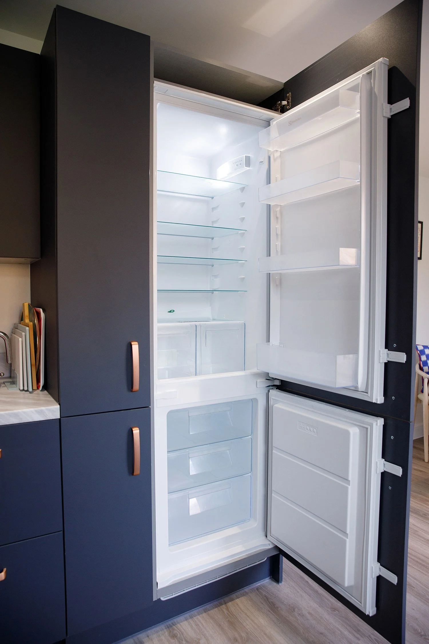 Empty modern refrigerator with the door open, inside shelves are clear, located in kitchen with dark cabinetry and wood flooring.