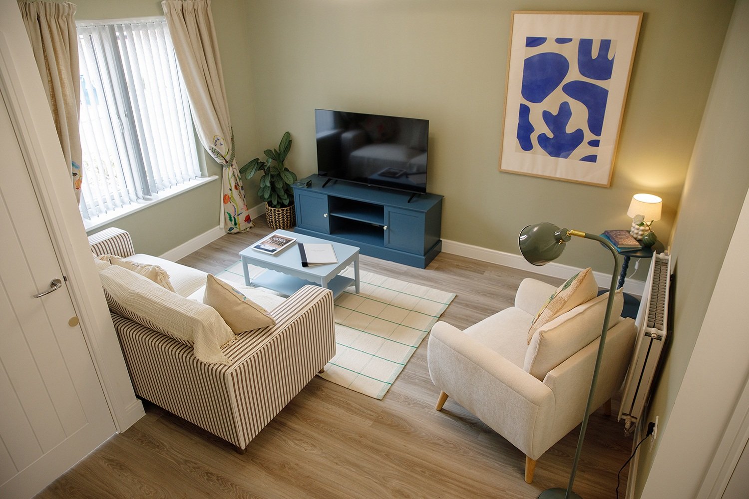 A cozy living room with a light green wall, a striped sofa with pillows, a white armchair, a blue TV stand with a flat-screen TV, a small coffee table, a patterned rug, and a large framed abstract art piece on the wall. There is a window with cream c