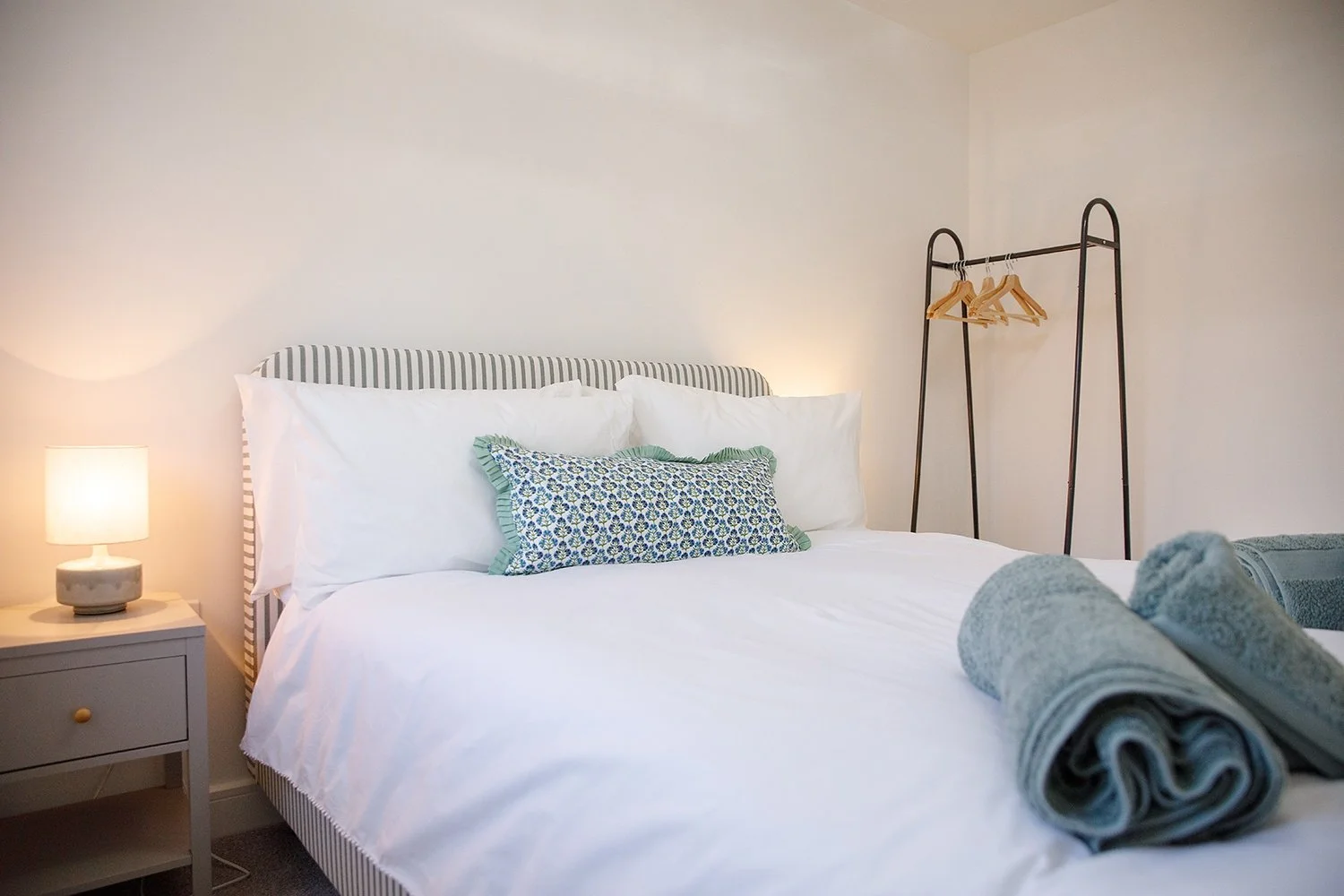 A neatly made bedroom with a bed, white bedding, decorative pillows, a nightstand with a lamp, and a clothing rack with hangers, in a minimalist style.