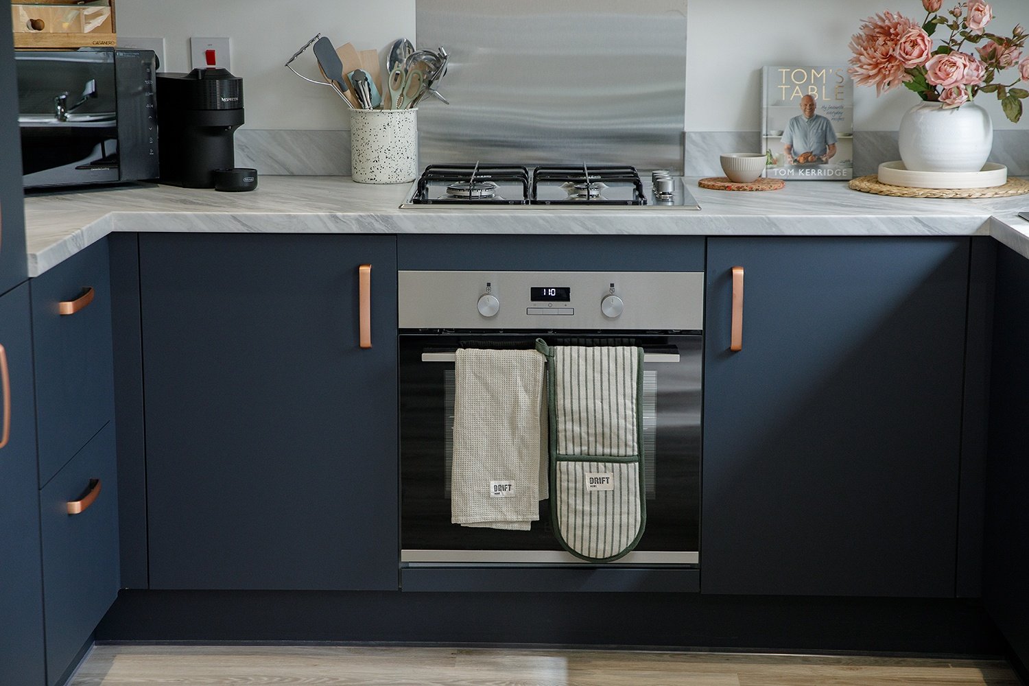 Modern kitchen with navy cabinets, stainless steel oven, gas stove, white marble countertop, pink flower vase, cookbook, and kitchen utensils