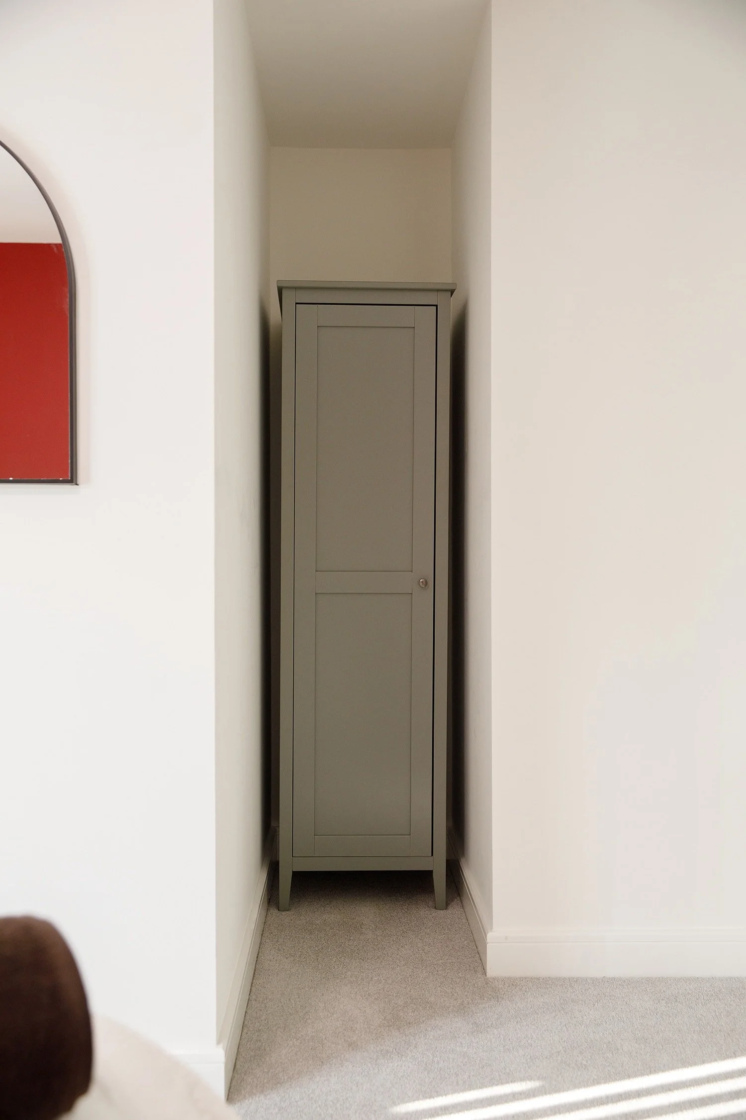 A tall, narrow, closed gray cabinet standing in a small alcove between two white walls with carpeted floor.