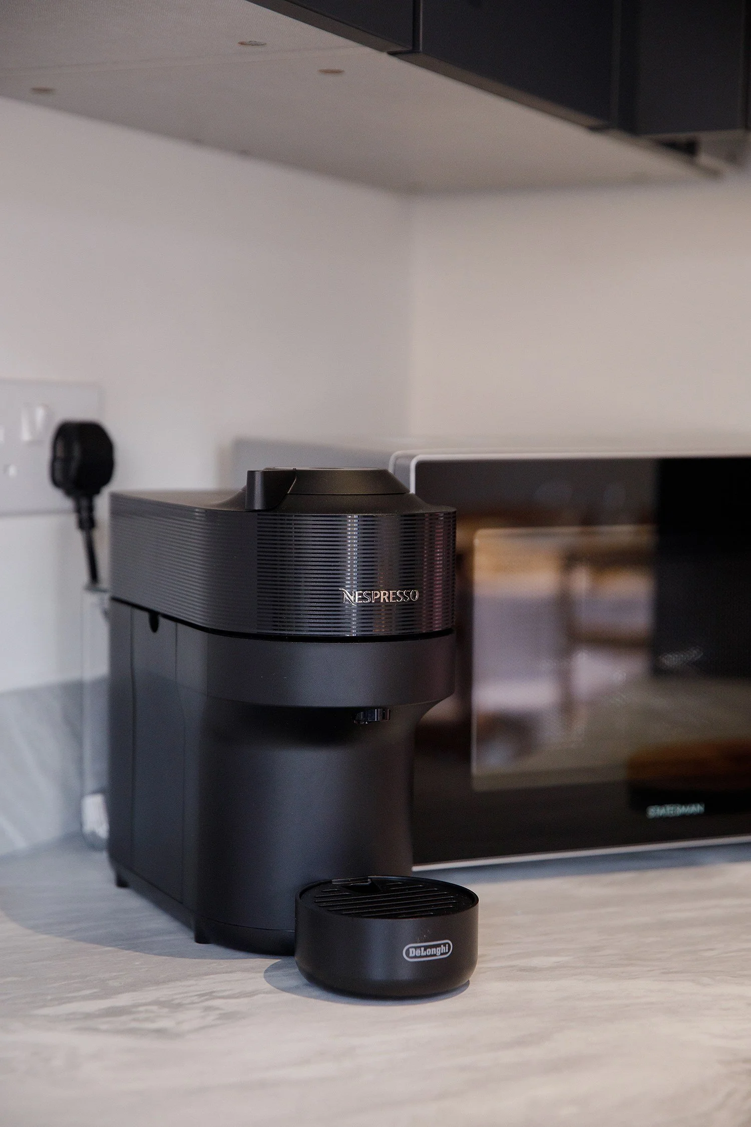 A Nespresso coffee machine on a kitchen countertop with a DeLonghi coffee capsule in front of it. In the background, a microwave oven and kitchen wall socket are visible.