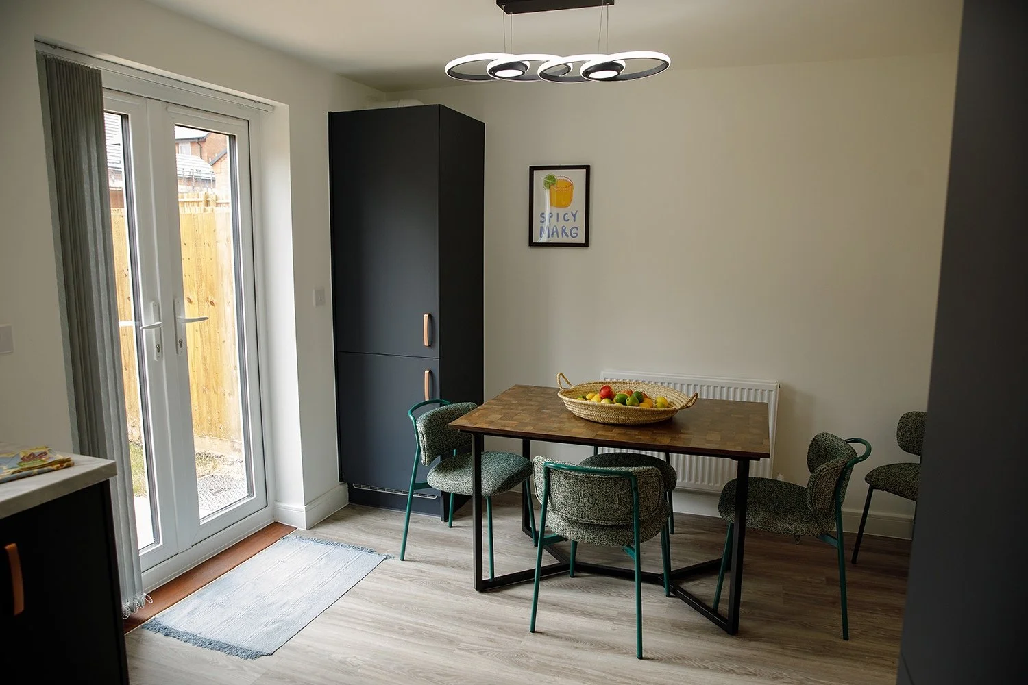 Modern dining room with a wooden table, four upholstered chairs, a black cabinet, a framed artwork, a basket of fruit, and glass doors leading outside.