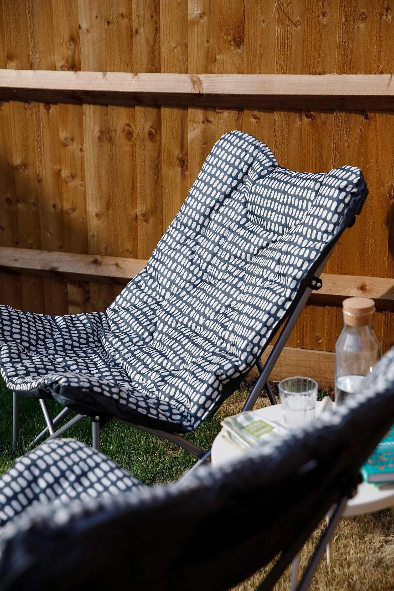 Garden scene with a black and white polka dot lounge chair in the foreground, a wooden fence in the background, a table with a bottle, glasses, and snacks.