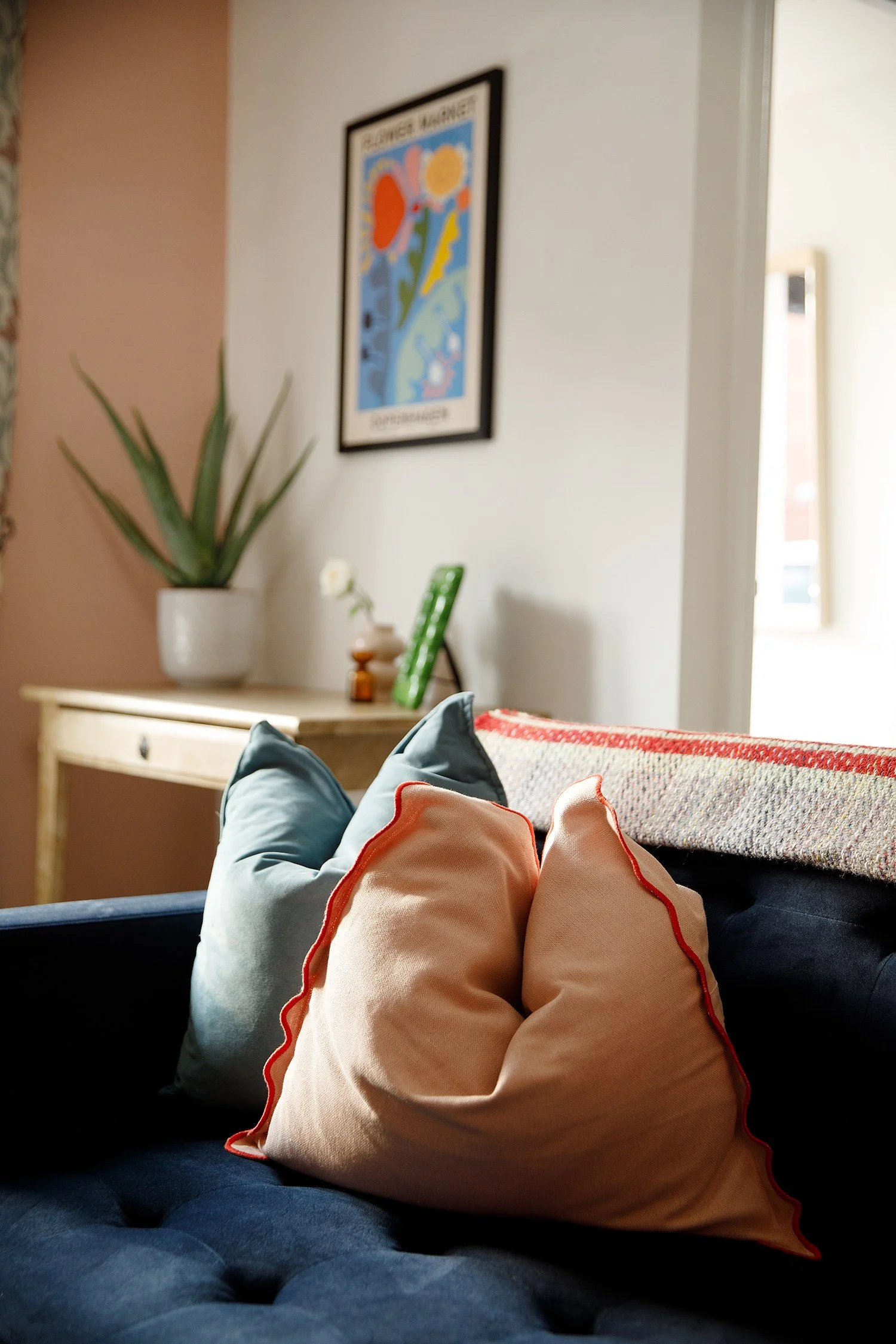 Interior of a living room or similar space, viewed from a person sitting on a dark blue couch. The image shows pillows on the couch, including a visible peach-colored pillow with red piping. In the background, a wooden console table holds a potted pl