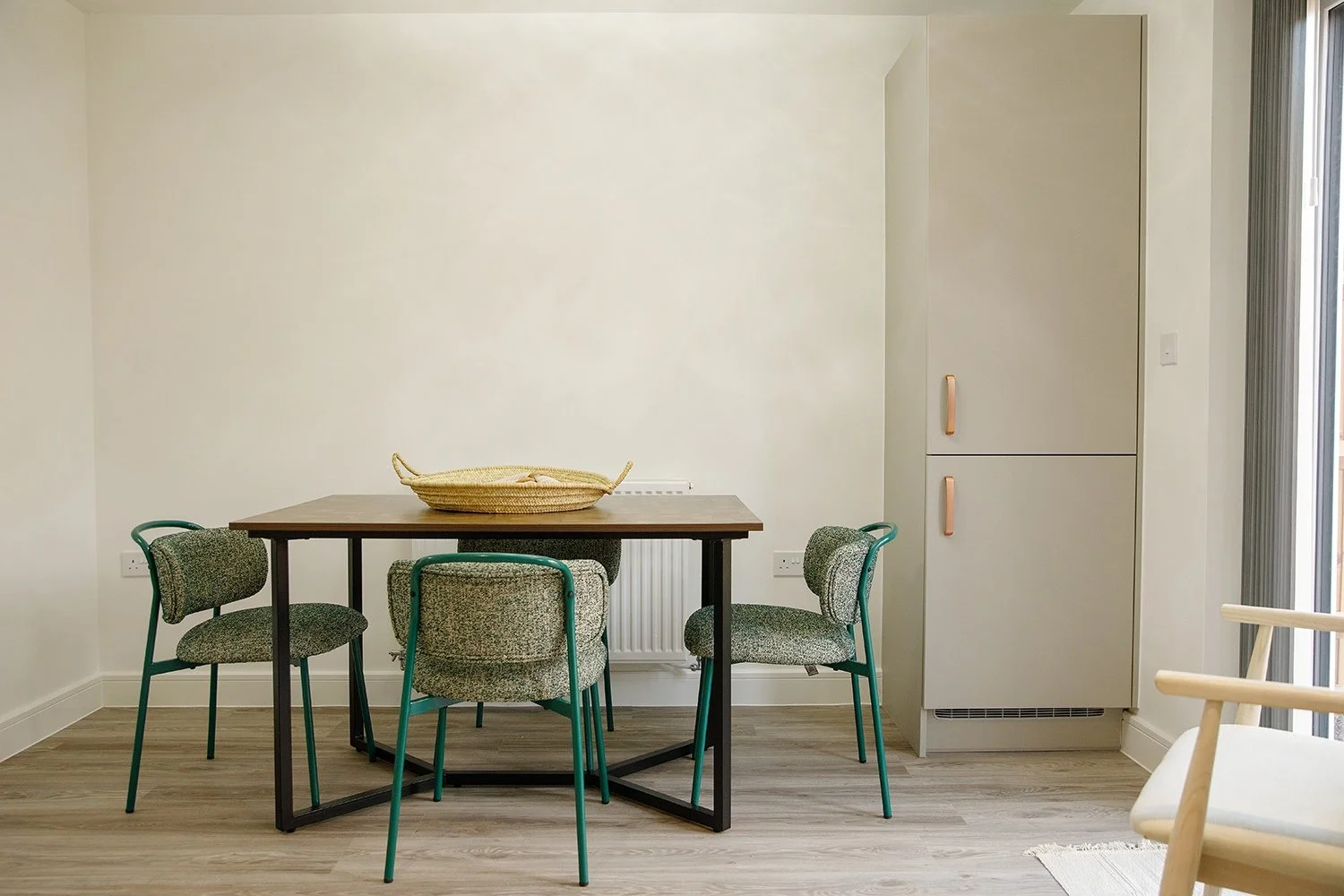 Dining area with a wooden table, four green and gray chairs, a woven basket on the table, and a tall white cabinet against a light-colored wall. A sliding glass door is on the right.