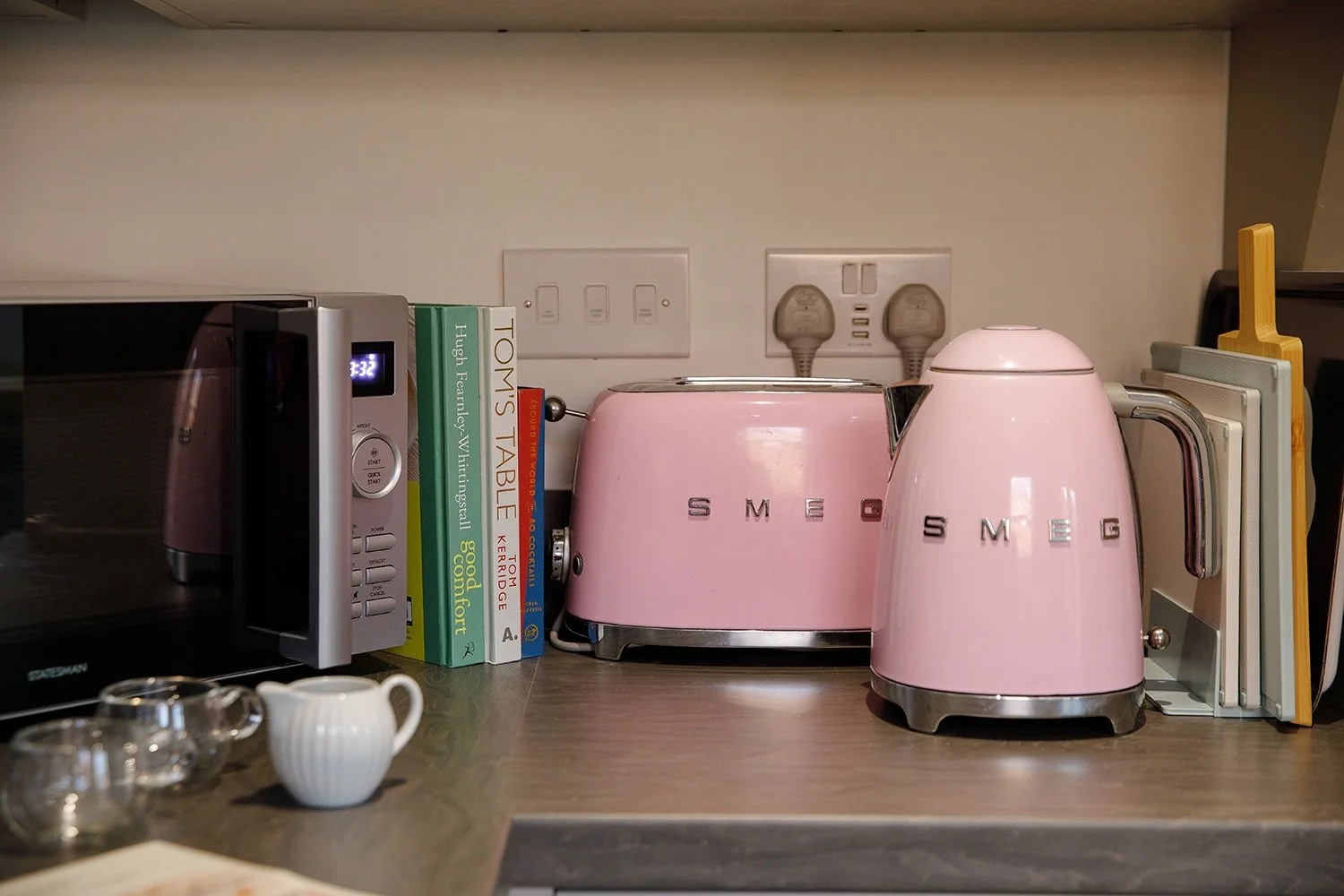 Pink toaster and kettle on kitchen countertop with cookbooks and small white pitcher, electric outlets in background.