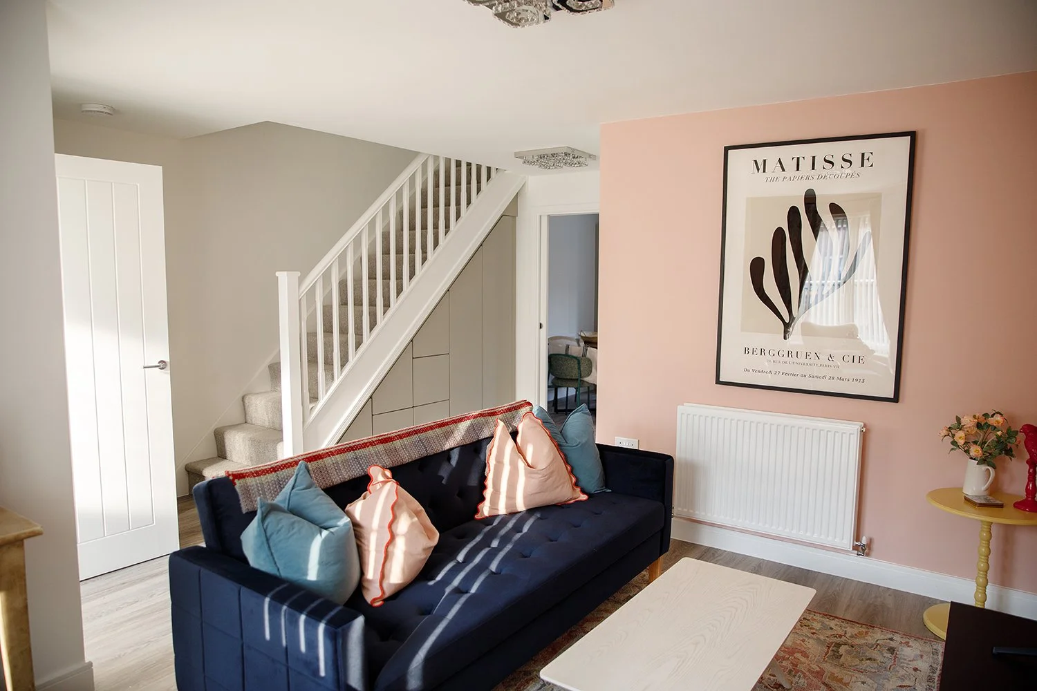 Living room with a navy sofa, colorful cushions, a wall with a pink accent, and a large framed art print. There's a small yellow side table with flowers and a radiator underneath the art. A staircase with carpeted steps and white railing leads upstairs.