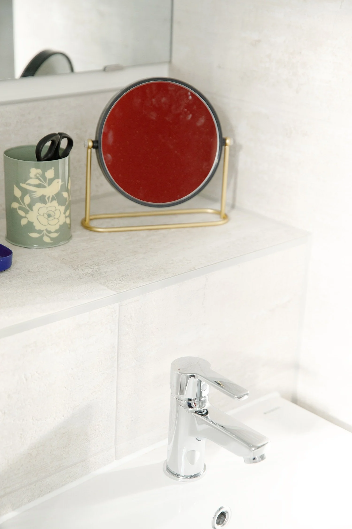 Kitchen sink with a silver faucet, a mirror, a green cup holding scissors with a floral pattern, and a small round mirror with a gold stand on a white countertop.