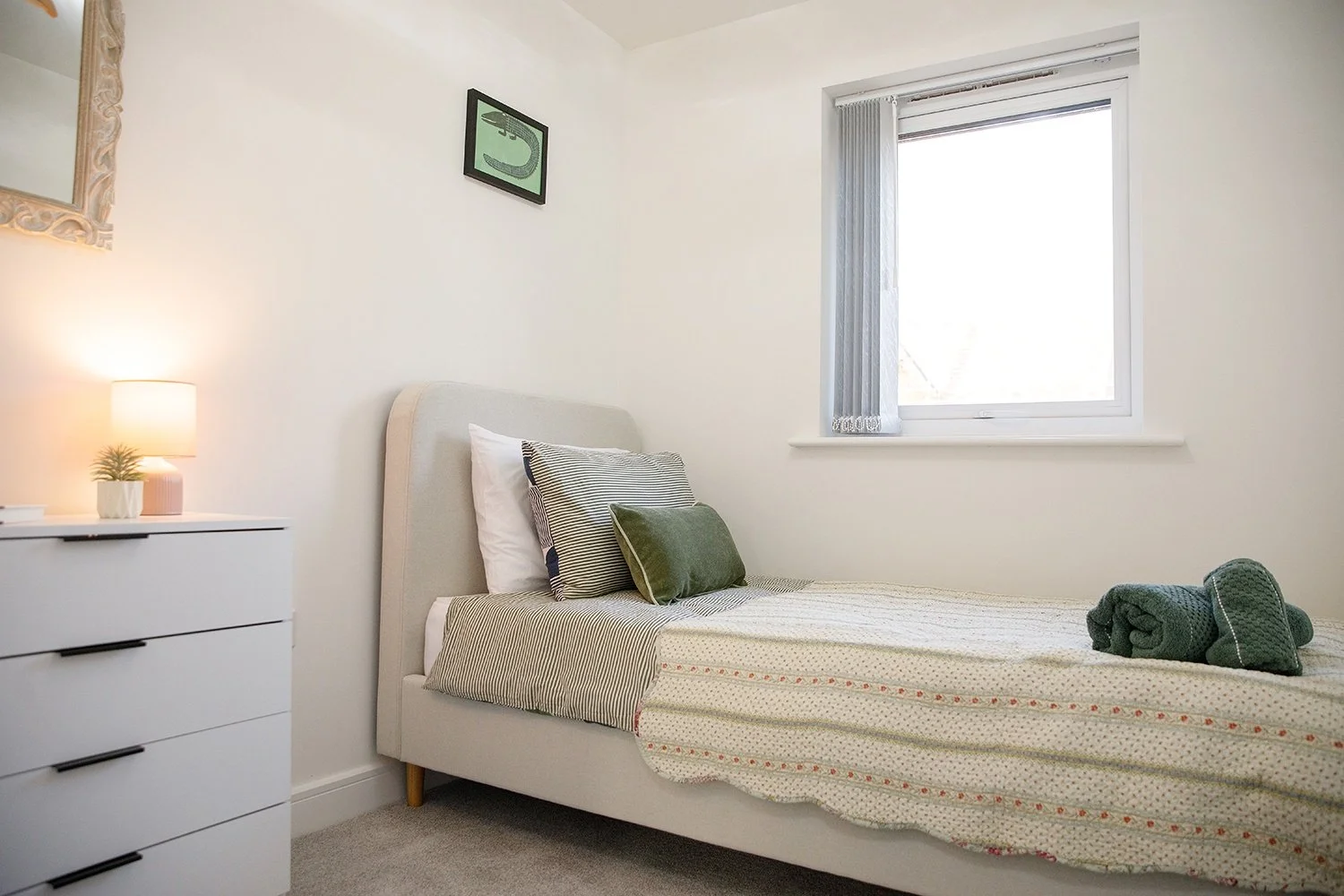 A neatly made single bed with a cream-colored headboard, striped pillows, and folded green towels at the foot. A white nightstand with a lamp and small plant is beside the bed. There is a window with vertical blinds and a small framed art piece above