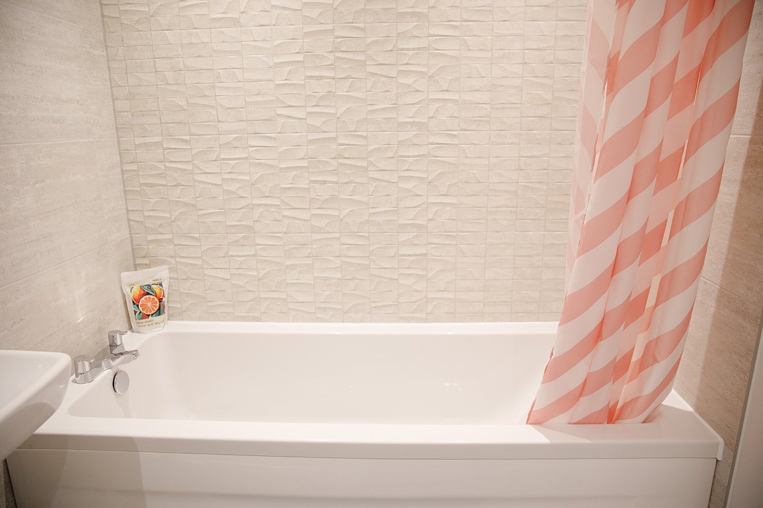 Bathroom with white bathtub, pink and white striped shower curtain, beige textured tile wall, and a small soap dispenser on the edge of the tub.