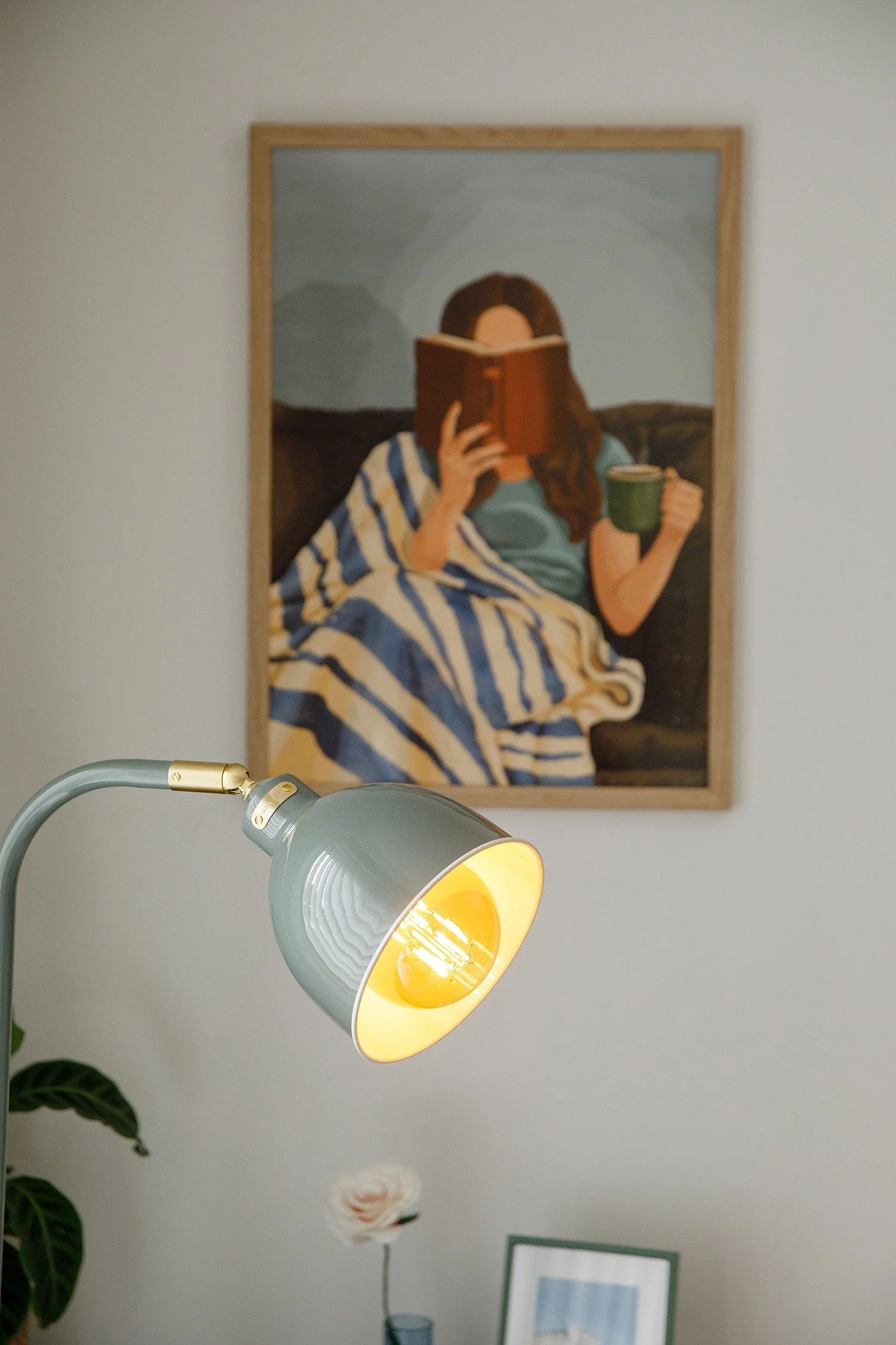 Painting of a woman reading a book while holding a mug, with a mountain landscape background, hung on a beige wall.