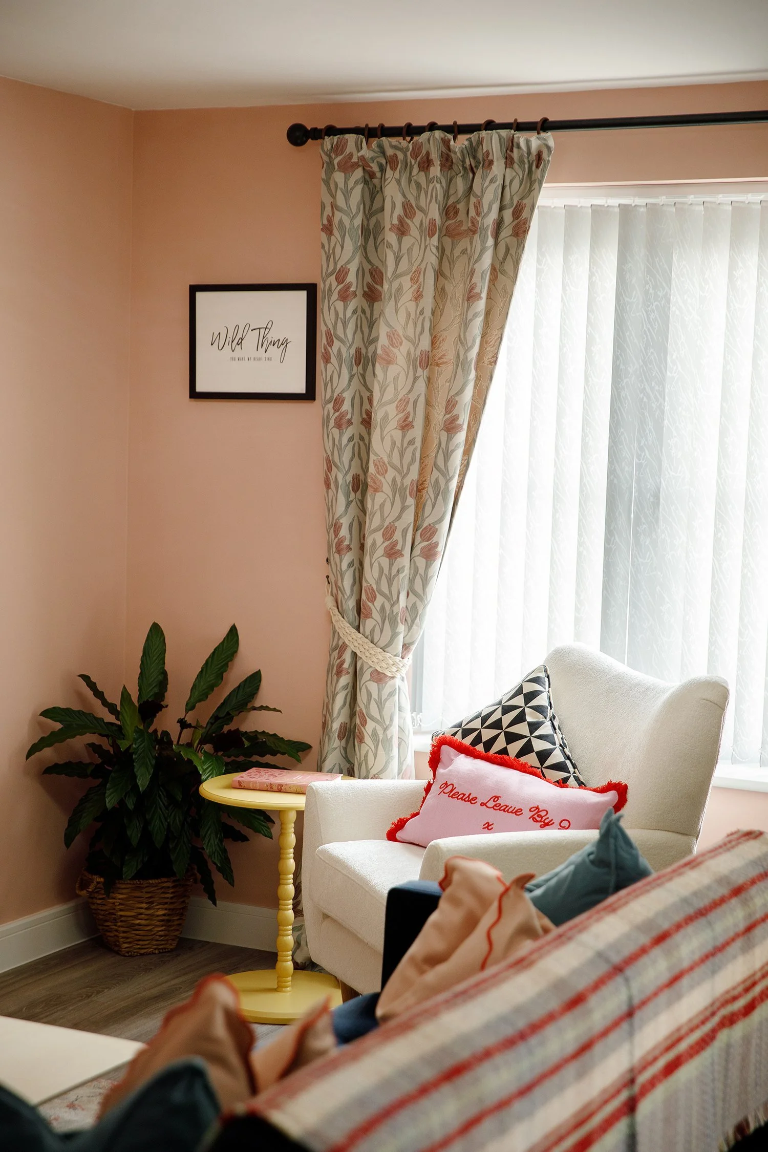 A cozy living room corner with a window dressed with floral-patterned curtains and vertical blinds. A white armchair with pillows, including one with red piping and embroidery, is positioned near a small yellow side table with books. A large potted p