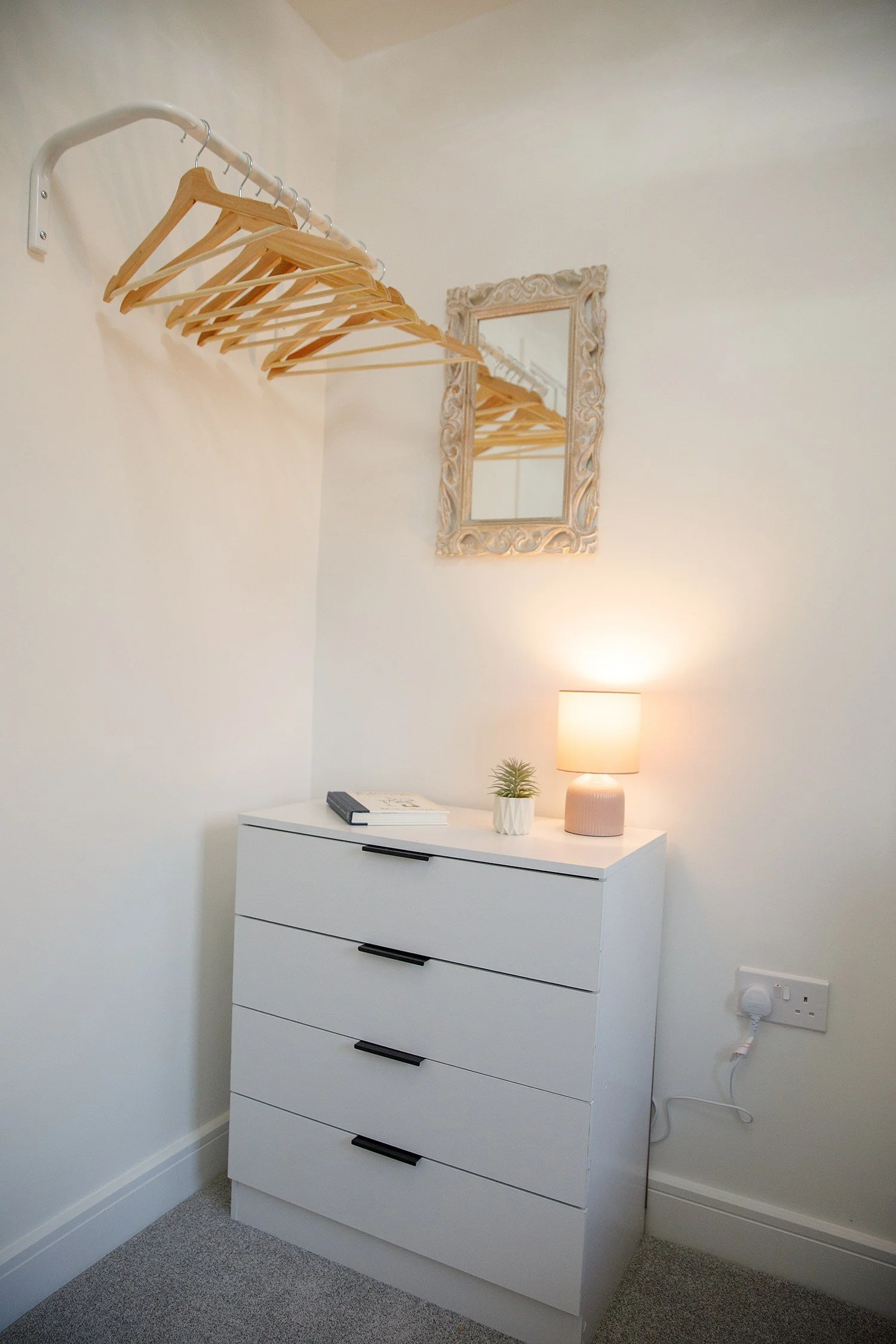 A white dresser with four drawers, a small potted plant, a table lamp, and a closed book on top, against a wall with a mirror, wooden hangers on a rack, and electrical outlets.