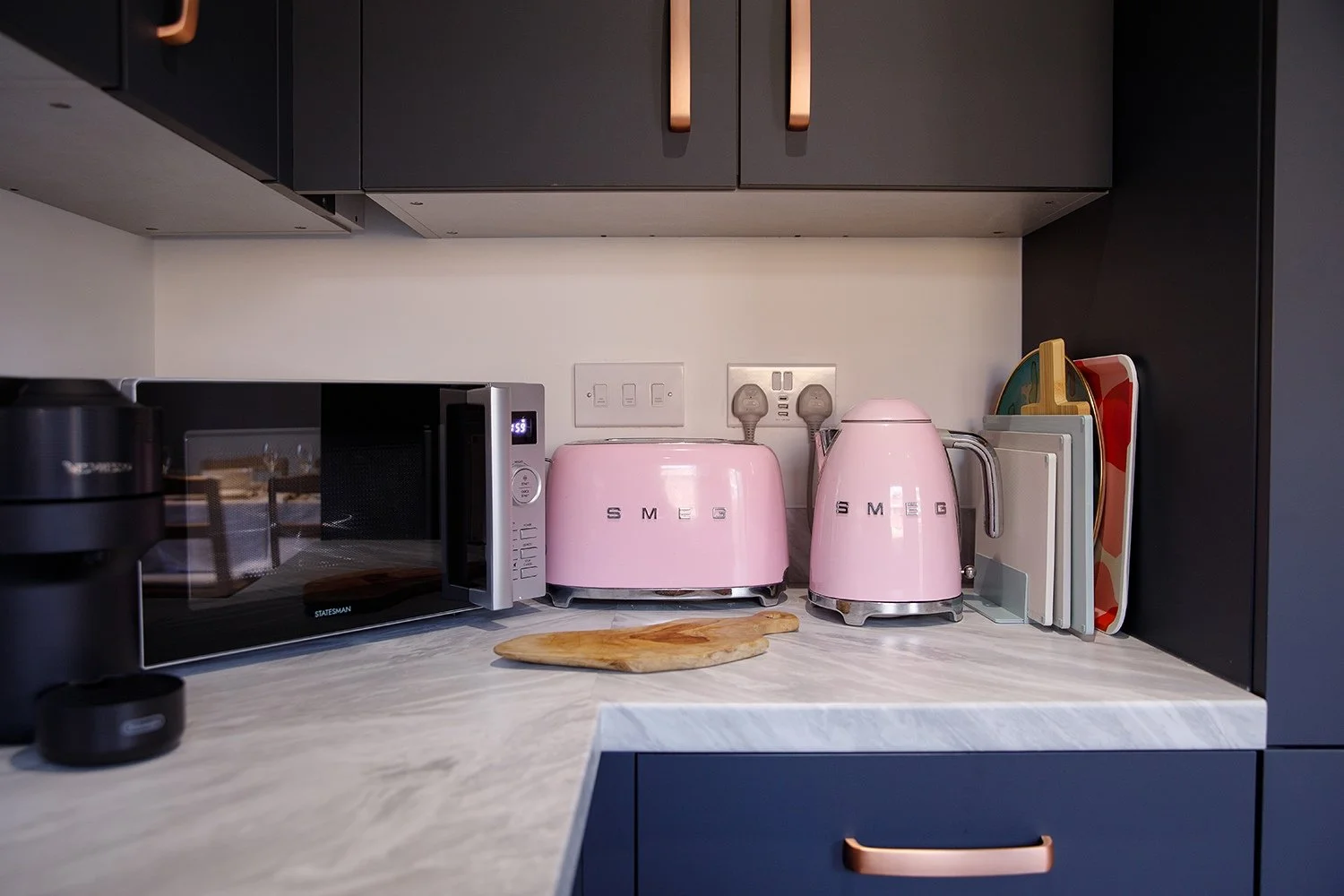 Kitchen countertop with a black microwave, a pink toaster, a pink kettle, and a knife block with colorful knives.