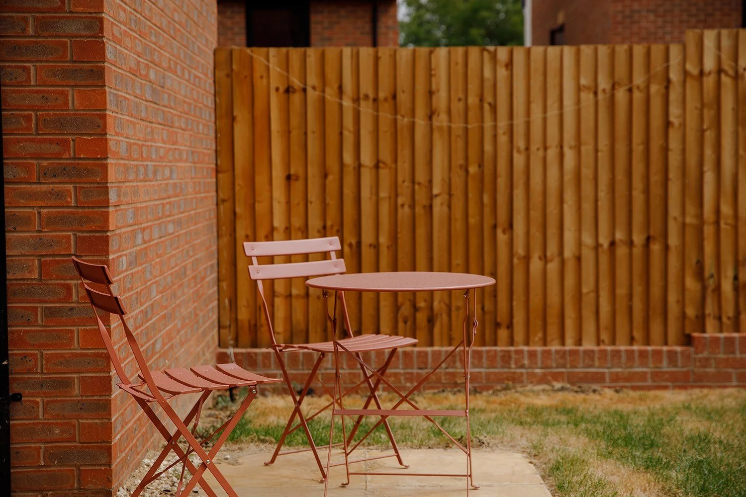 Pink metal patio table and chair set on a concrete slab in a backyard with a brick wall and wooden fence.