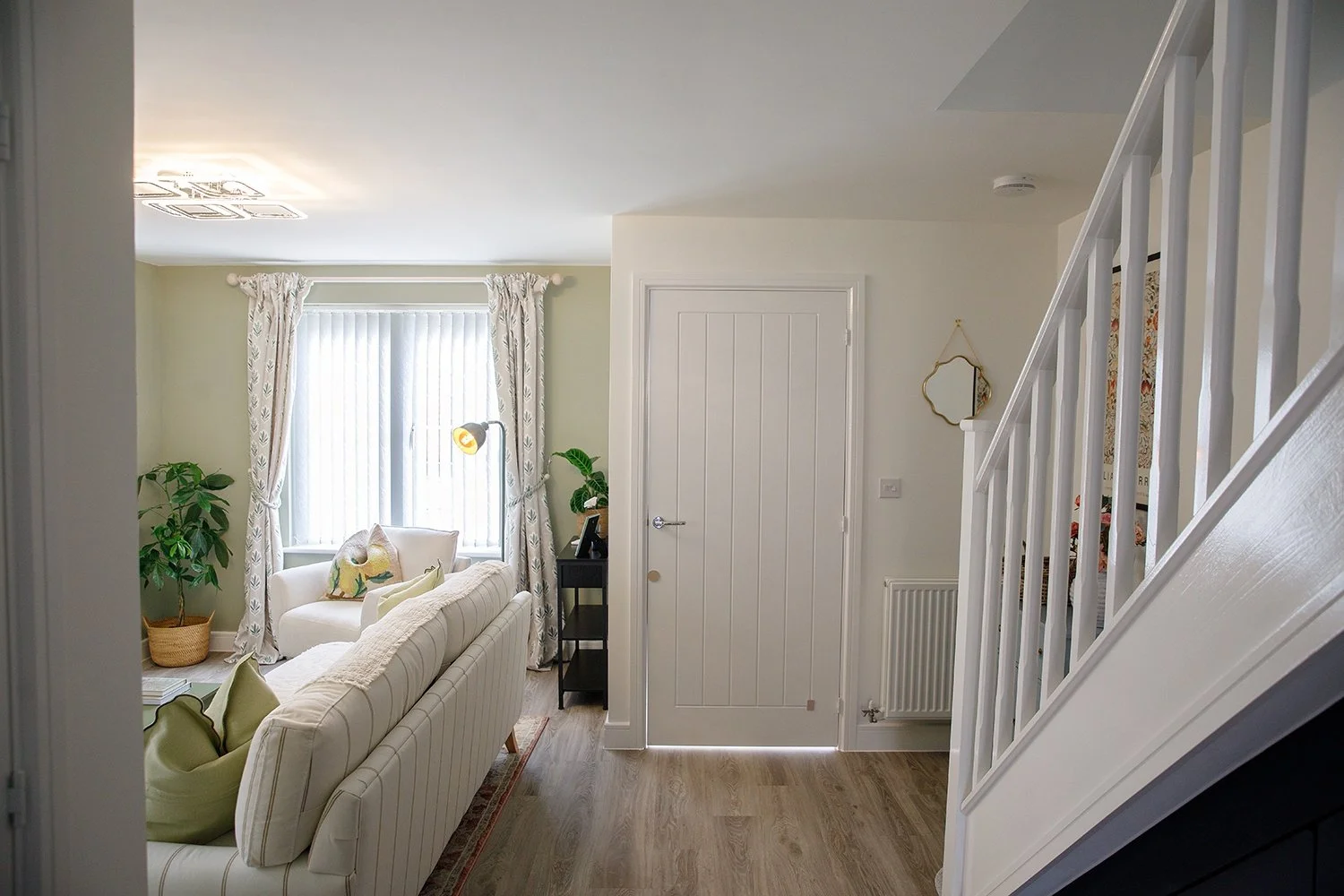 Interior view of a living room with a white armchair, green plants, a window with patterned curtains, and stairs on the right.