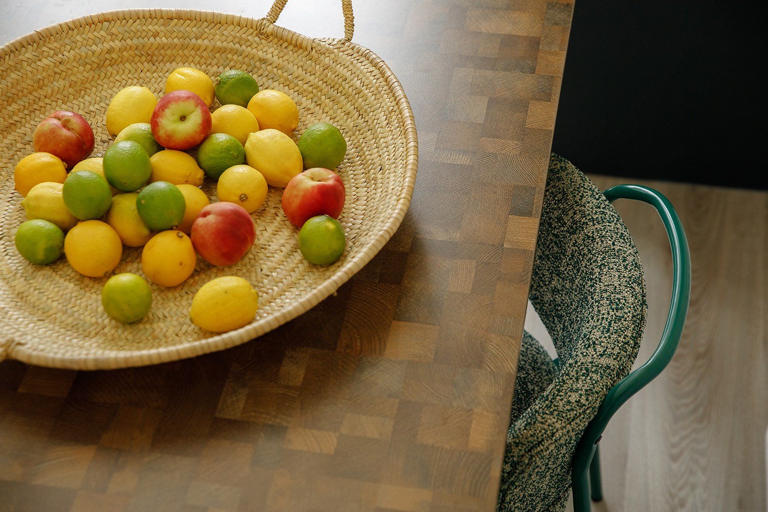 A woven basket filled with lemons, limes, and apples on a wooden table next to a patterned chair.