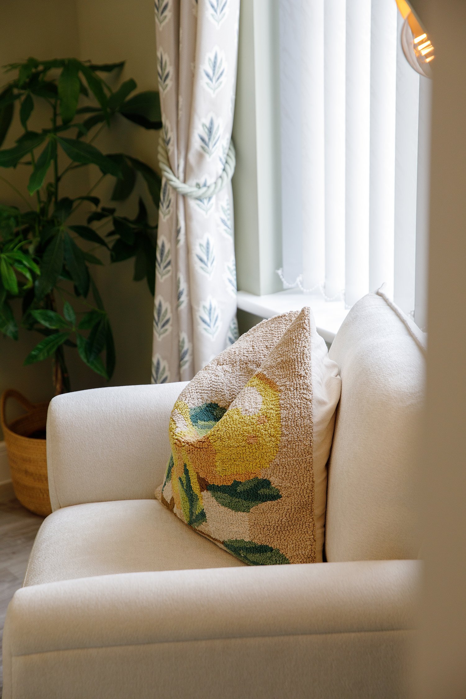 A cozy living room corner with a white armchair, a colorful floral pillow, a large potted plant with broad green leaves, white curtains with a leaf pattern tied with a rope, and a window with sheer white blinds letting in natural light.