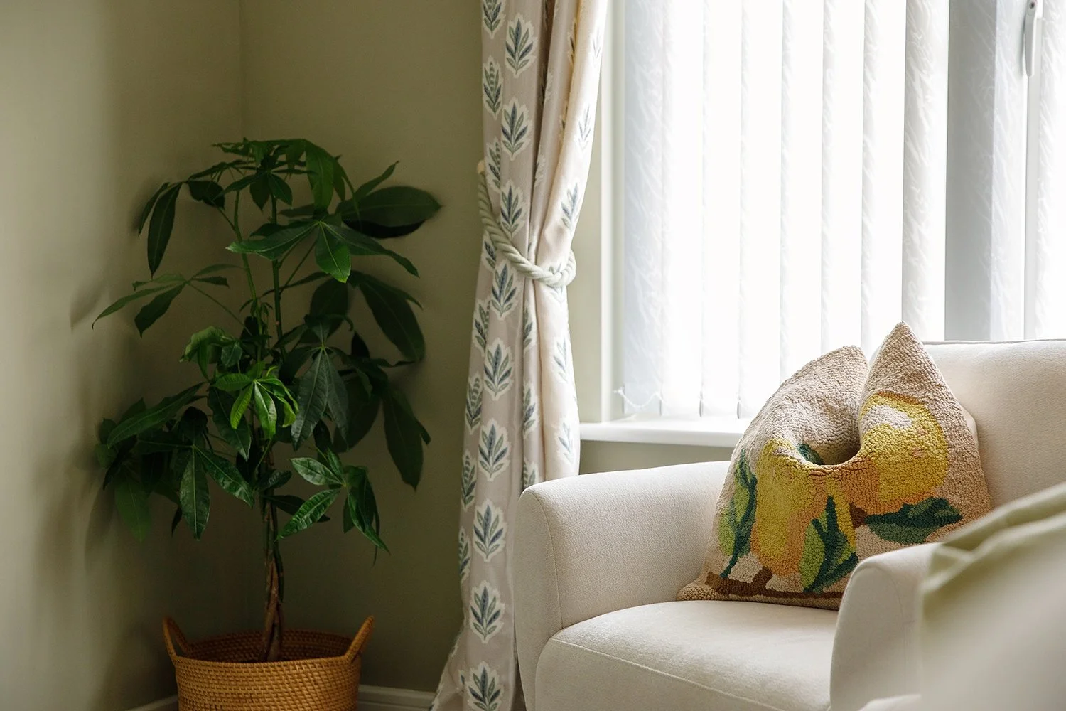 A cozy corner with a beige sofa, a decorative pillow with a floral pattern, a large leafy houseplant in a woven basket, and a window with sheer white curtains and patterned drapes tied back.