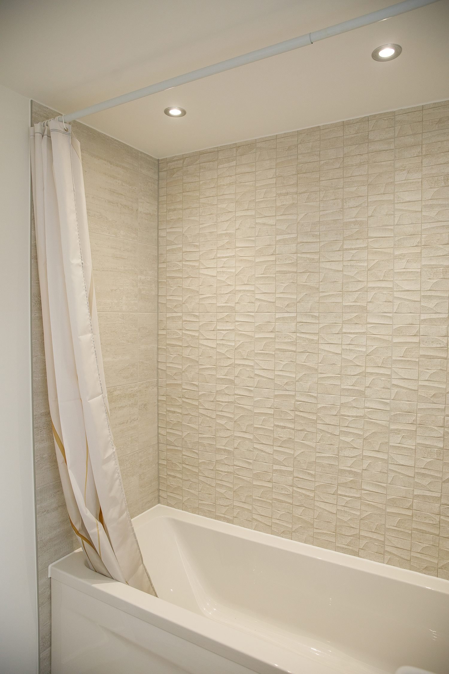A bathroom shower with beige textured tiles on the wall, a white bathtub, a white shower curtain with gold trim, and two recessed ceiling lights.