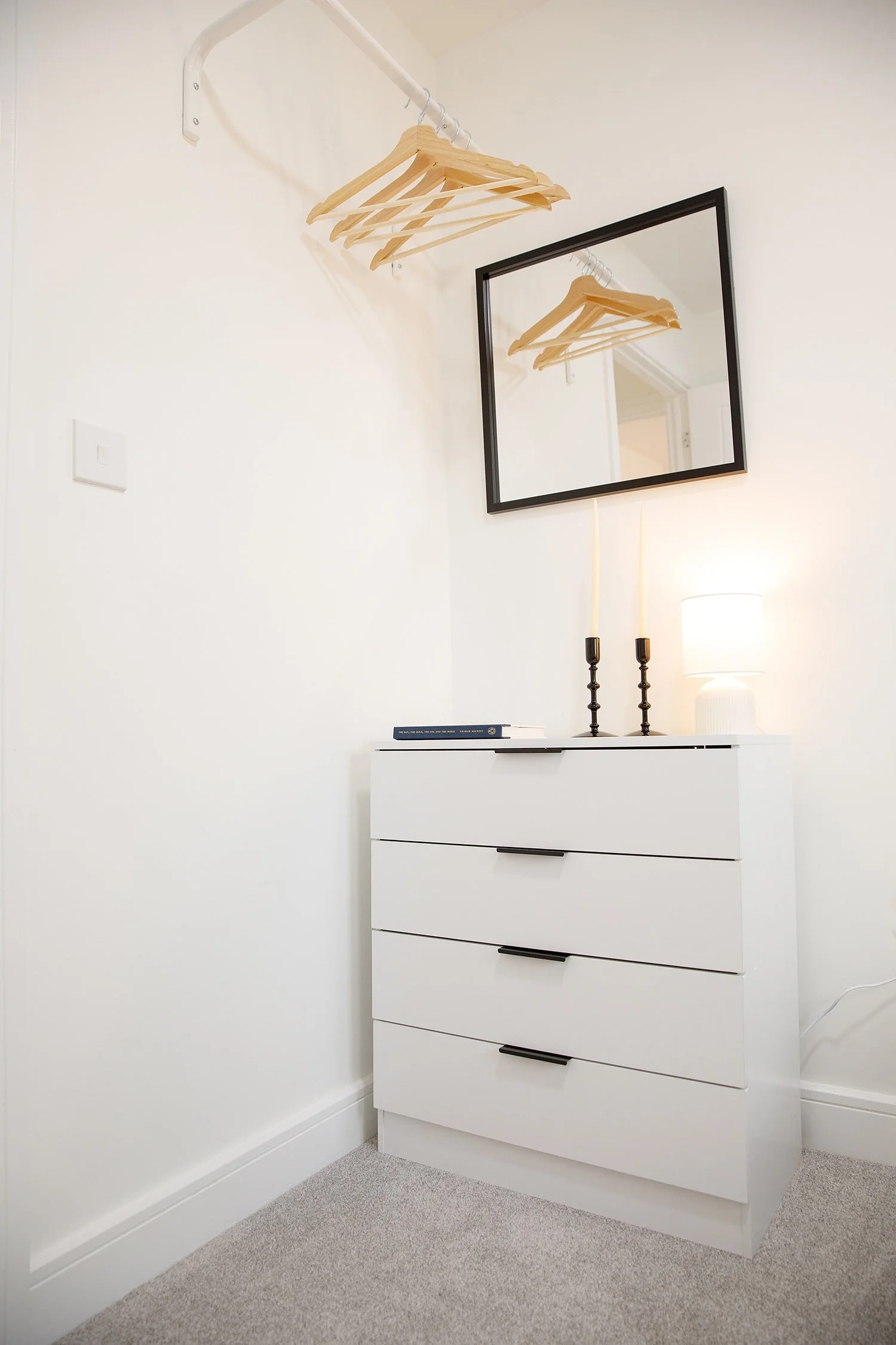 A white dresser with five drawers stands against a white wall in a room. On top of the dresser, there are two black candle holders with candles, a white lamp, and a book. A large rectangular mirror hangs on the wall above the dresser, reflecting the 