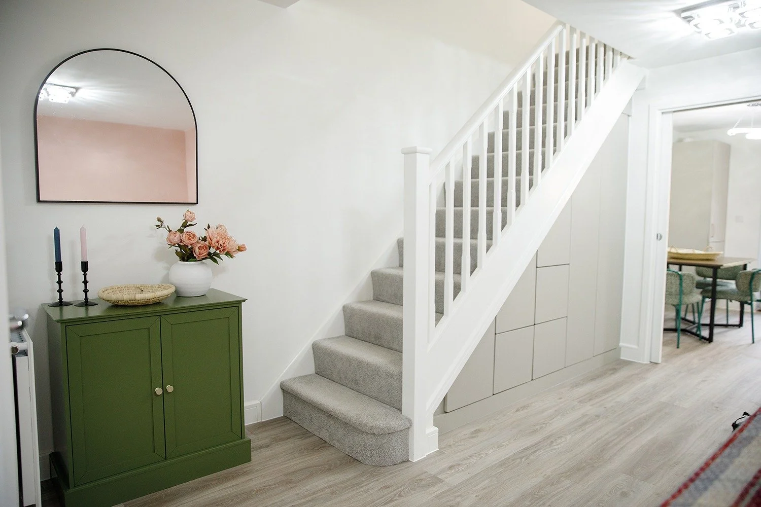 Interior view of a home with a staircase, a green cabinet with a flower vase and candles, and a mirror on the wall.