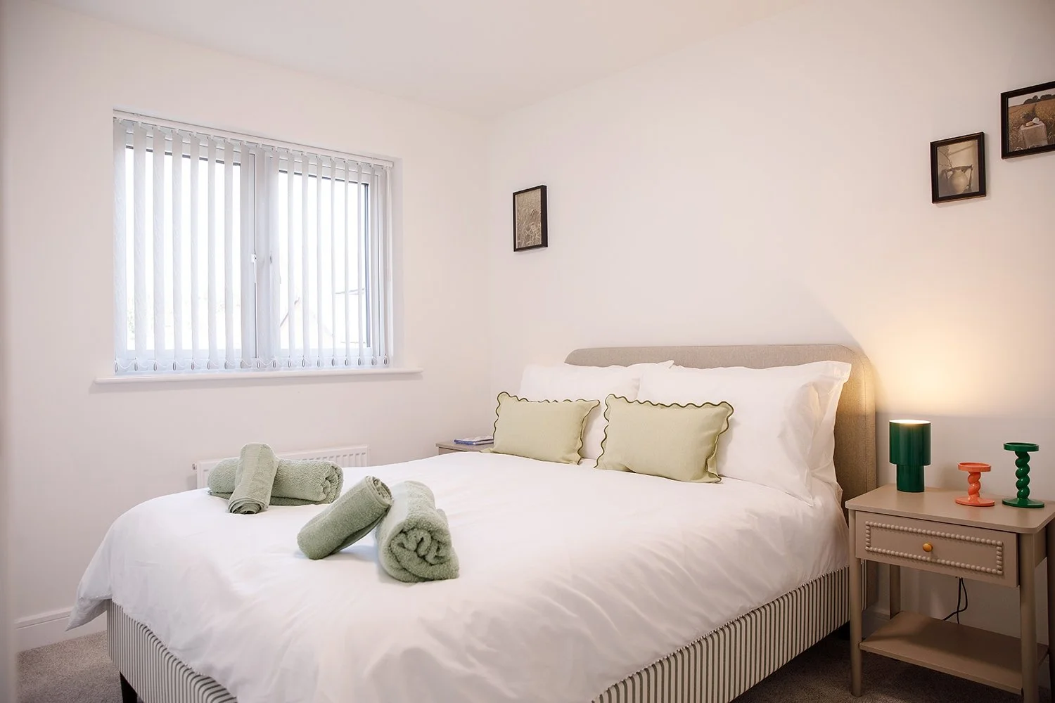 A neatly made bed with three rolled towels on top, two green pillows, and white bedding. There is a bedside table with decorative lamps and a radiator below a window with vertical blinds. The wall is decorated with three framed pictures.