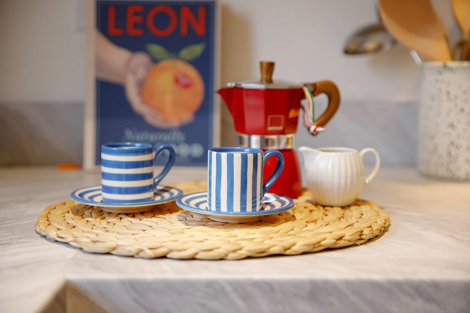 Two blue and white striped coffee cups and saucers, a small white creamer pitcher, sitting on a round woven placemat on a countertop, with a red moka pot and a poster in the background.